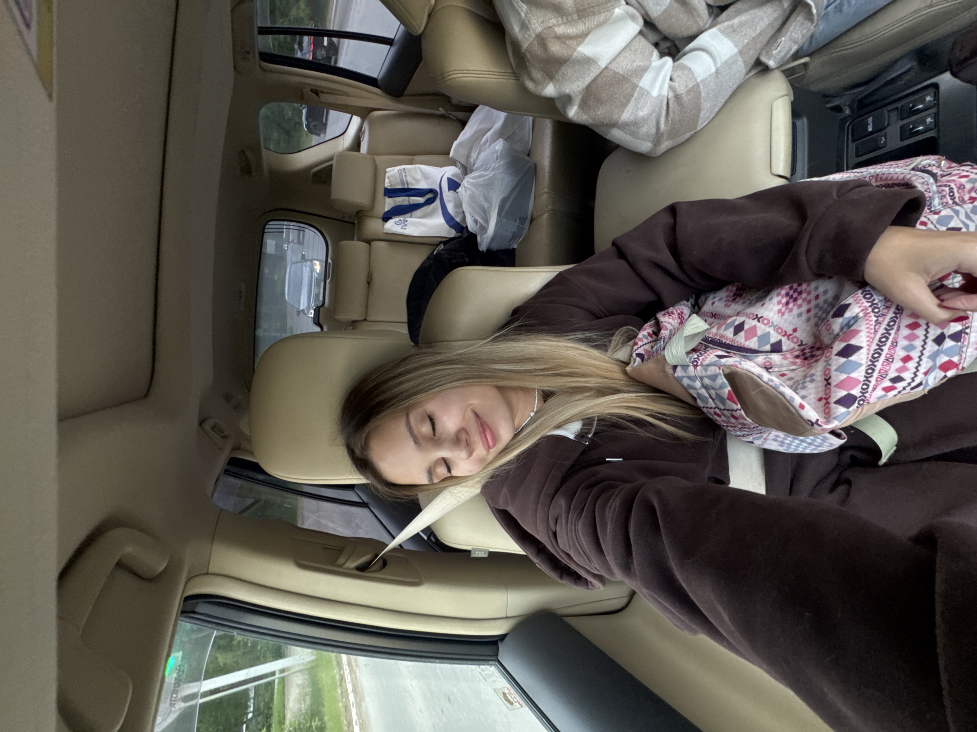 A young woman with blonde hair and closed eyes is resting in the passenger seat of a car, holding a bag with a patterned design. She is wearing a brown hooded sweatshirt. The car's interior is visible, with tan leather seats and a beige headliner. Bags and other items are seen in the back seat. The view from the car window shows a road and trees, suggesting an outdoor environment, potentially within the city of Chaikovsky, Russia.