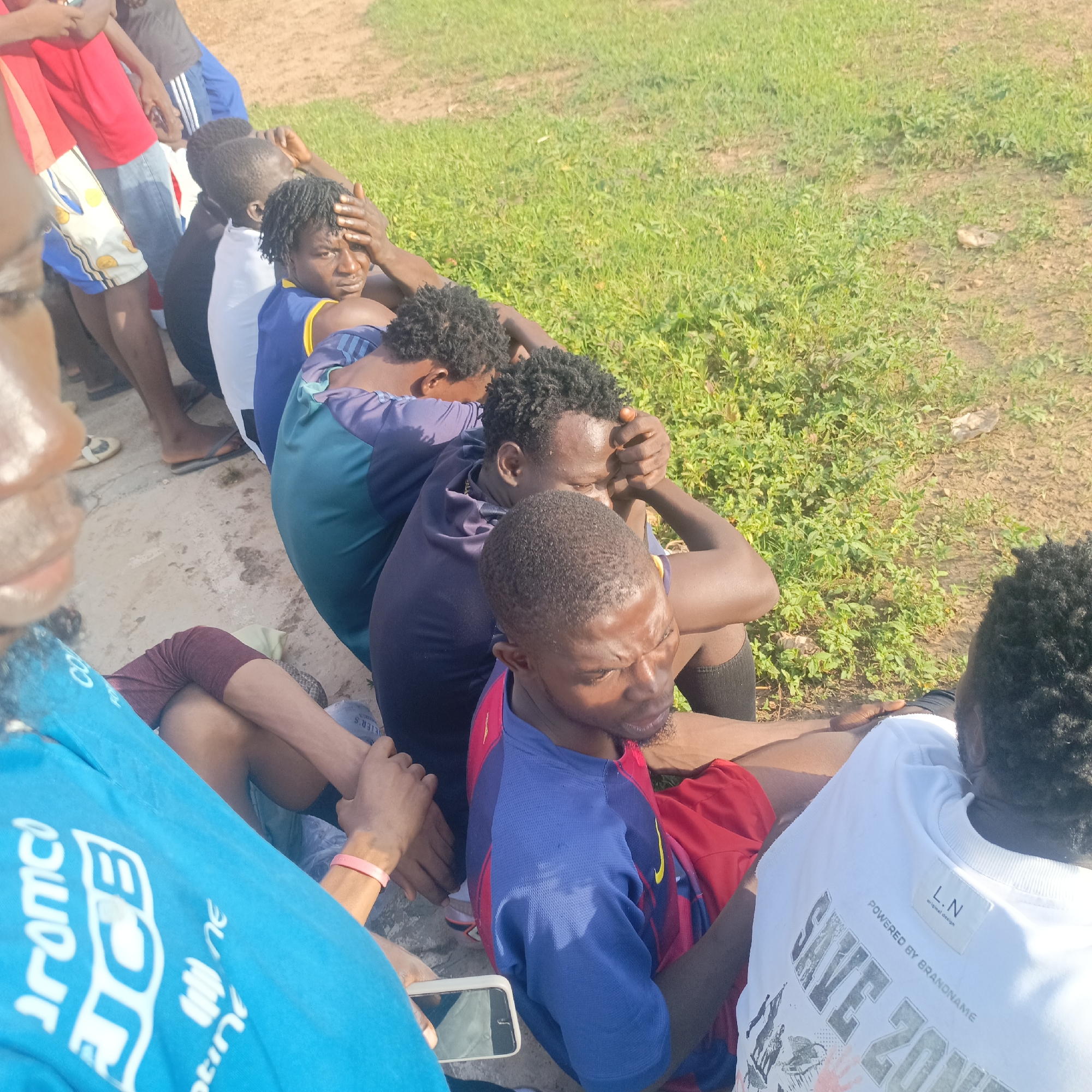 In Akwanga, Nigeria, a group of young Black men are seen sitting in a line on a concrete surface adjacent to a grassy area. The men are wearing casual athletic clothing, including t-shirts and shorts. Several individuals have their hands on their heads, with expressions that could suggest fatigue, distress, or contemplation. One person in the foreground, wearing a blue t-shirt with white lettering, is holding a smartphone. The overall impression is of a group taking a break or resting outdoors, possibly after physical activity. The environment appears to be an open, unpaved area with sparse vegetation.