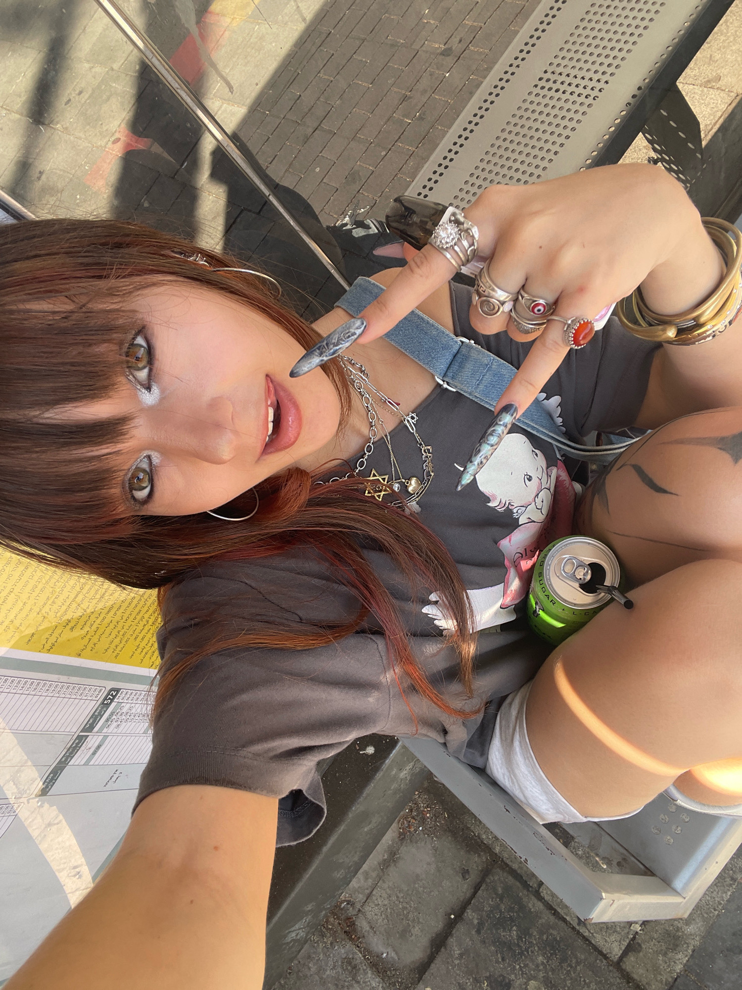 This is a high-angle, slightly over-the-shoulder selfie of a young woman sitting outdoors on a metal bench, possibly at a bus stop. The woman has long, reddish-brown hair with bangs and expressive eyes with noticeable eye makeup. She is wearing a dark t-shirt with a cartoon graphic and denim suspenders. She has long, decorative fingernails, multiple rings on her fingers, and several bracelets on her wrist.
In her right hand, she holds a can of "SUGA CA" soda with a black straw. A tattoo is partially visible on her upper thigh. The background shows a paved area and sunlight casting shadows, suggesting it is daytime, likely sunny. A yellow sign with Arabic and Hebrew text, appearing to be a bus schedule, is visible in the lower left corner. The text includes route numbers like "572" and destination names like "Jerusalem." The woman has a relaxed, somewhat playful expression, with her mouth slightly open. The scene appears casual and urban.