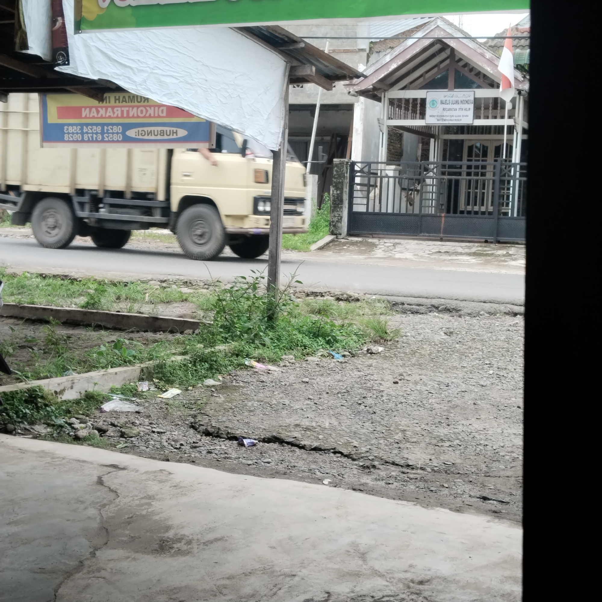 An outdoor scene in Talun Kenas, Indonesia shows a cream-colored truck driving on a paved road. A banner is attached to the side of the truck, displaying text in a foreign language and numbers. To the right of the truck is a building with a pitched roof, a metal gate, and an Indonesian flag. The foreground of the image features a cracked concrete surface with vegetation and scattered debris.
