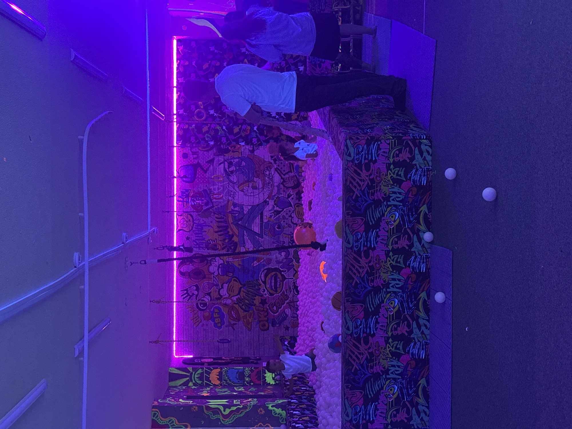 In this indoor scene bathed in vibrant purple and pink neon light, a ball pit dominates the right side of the frame, filled with countless white balls. On the left, a wall is adorned with colorful graffiti-style artwork and what appear to be stylized cartoon characters.
Two people are visible. In the foreground, a person in a white t-shirt and dark pants is partially visible, appearing to be interacting with the ball pit or its surroundings. Further back, another person is seen from the shoulders up, wearing a light-colored shirt.
The overall atmosphere is energetic and playful, likely an entertainment venue such as an arcade, trampoline park, or indoor playground. The neon lighting suggests the activity is taking place in the evening or a dimly lit indoor environment. There are no clear indications of time of day, weather, or specific geographic location beyond it being an indoor setting. The graffiti art, while colorful and detailed, does not present discernible text that can be easily read. A few white ping pong balls are scattered on the floor in the upper right corner of the image.