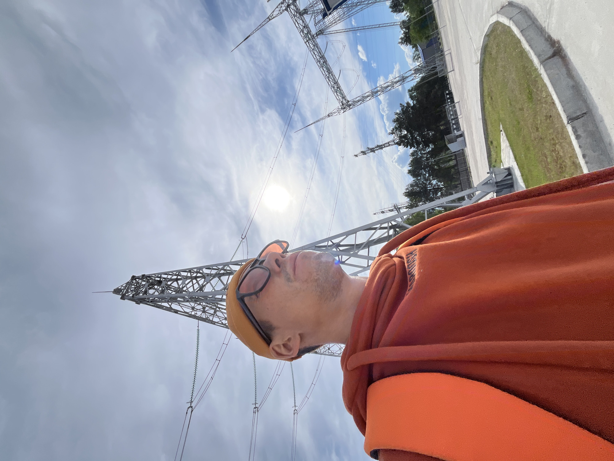 A low-angle outdoor portrait features a man gazing upwards towards a bright, partially cloudy sky filled with large metal electricity transmission towers and numerous power lines. The man, visible in profile from the chest up, wears an orange hooded sweatshirt, a yellow beanie, and dark-rimmed glasses. His head is tilted back as he observes the industrial structures and the sky above.
The sun appears as a strong glare point through the clouds in the lower-left sky, indicating bright daylight. Several tall lattice-style pylons dominate the background, supporting multiple power lines with visible greenish-blue insulators. A paved area with a curved grassy median is visible in the distance to the upper-right, alongside some distant trees. On the man's orange hoodie, a faint text reading "HOOD" is discernible. The scene captures an industrial or utility setting under a clear, sunny sky with scattered clouds, set in Chaikovsky, Russia.