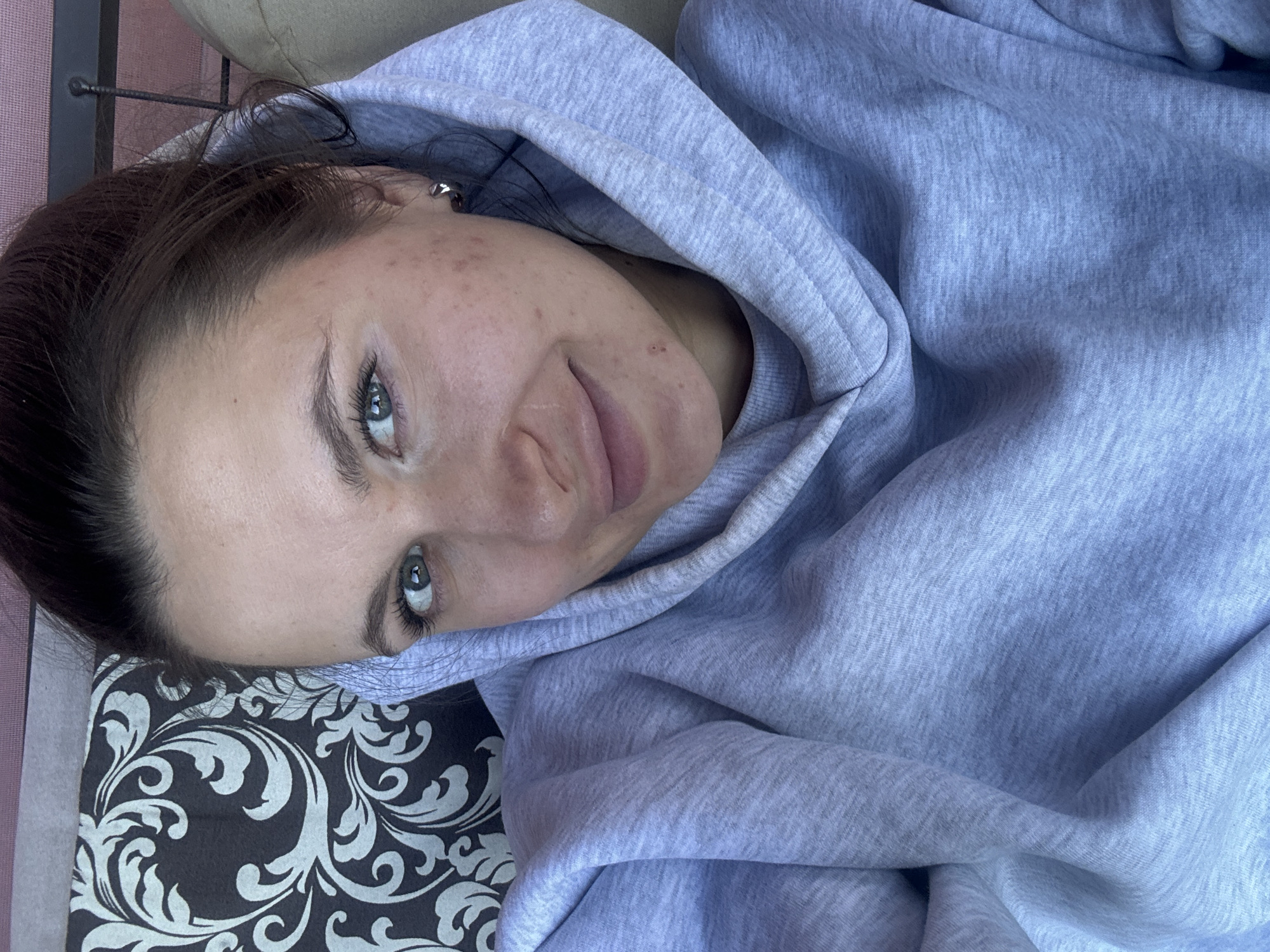 A woman with fair skin and green eyes is lying down, looking at the camera. She is wearing a grey hooded sweatshirt. Her hair is dark brown and pulled back. She has a silver earring. Her face shows some skin imperfections. The woman is resting her head on a patterned black and white cushion, which is placed on a surface that has pink stripes. It is not possible to confirm whether the location is in the city of Vysokinichi, Russia, based on the information in the image.