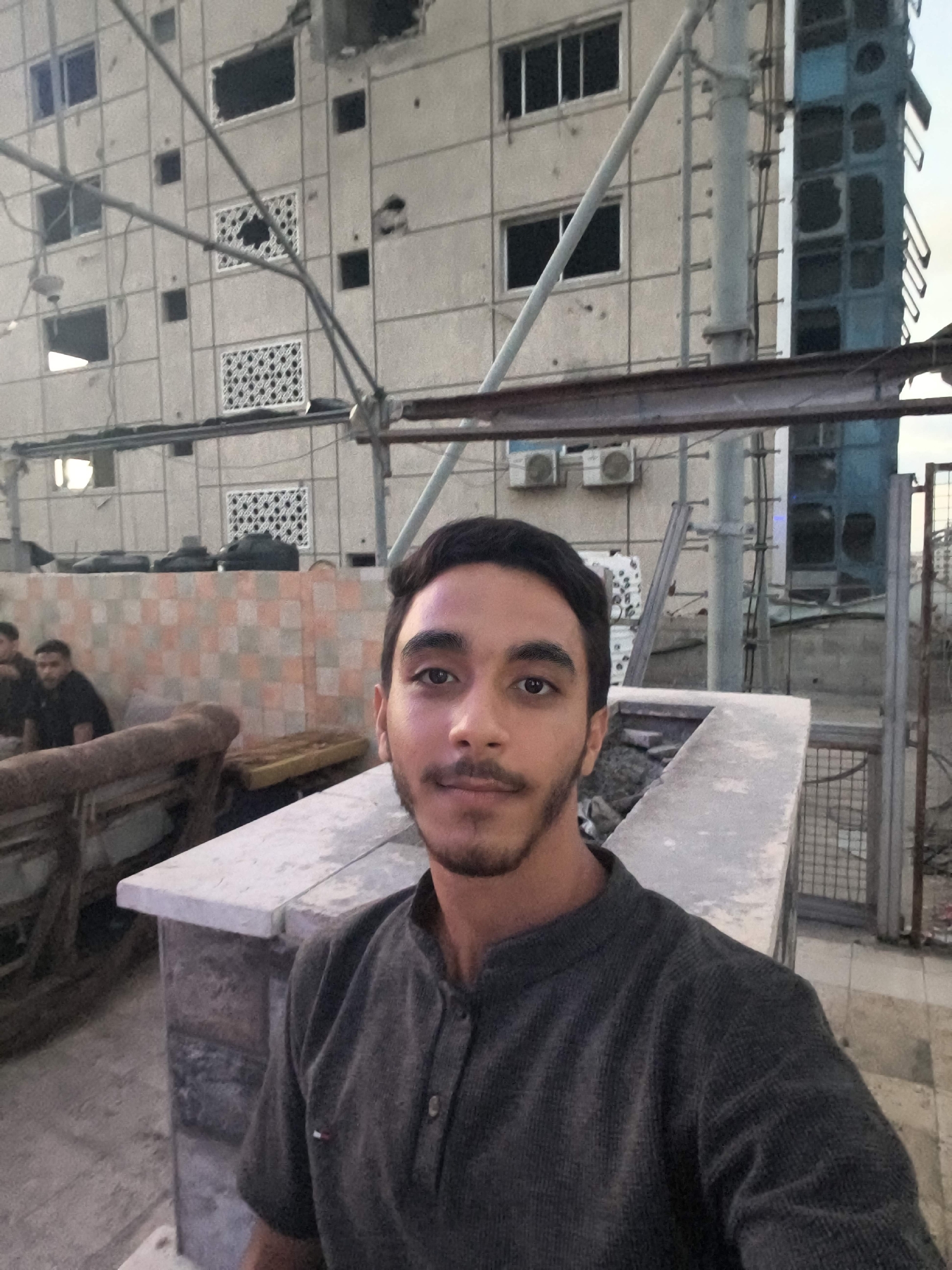 In Gaza City, Palestinian Territories, a young man with dark hair and a beard takes a selfie in the foreground. He is wearing a dark grey corduroy shirt. Behind him, to the left, two other men are seated. The background reveals a damaged, multi-story building with numerous holes and missing windows, suggesting destruction. Scaffolding and construction materials are visible, indicating ongoing or interrupted repair work. An air conditioning unit is mounted on the wall of the building. The scene appears to be outdoors, possibly a rooftop or elevated area, with a concrete structure in the middle ground. The overall impression is one of urban damage and resilience.