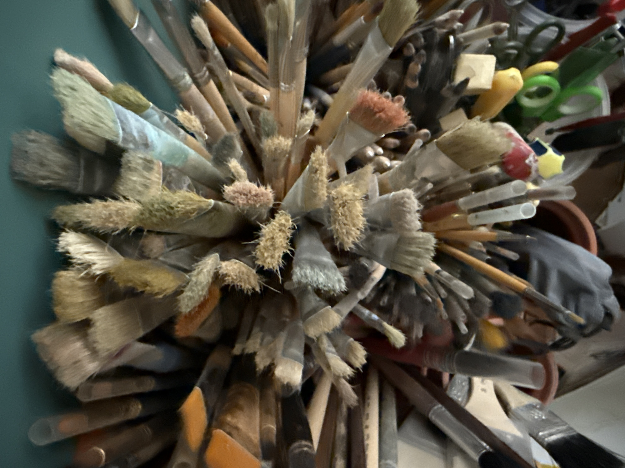 The image shows a close-up, overhead view of a large collection of art brushes, with their bristles splayed outwards. The brushes vary in size, color, and bristle type, with some appearing worn and others newer. They are bundled together, with their handles mostly obscured. Some of the brushes appear to be made of wood or plastic, with ferrules made of metal. In the background, the teal color of a wall or ceiling is visible. The scene is consistent with an artist's studio or art supply storage area. There is no indication of the city of Votkinsk, Russia in the image.