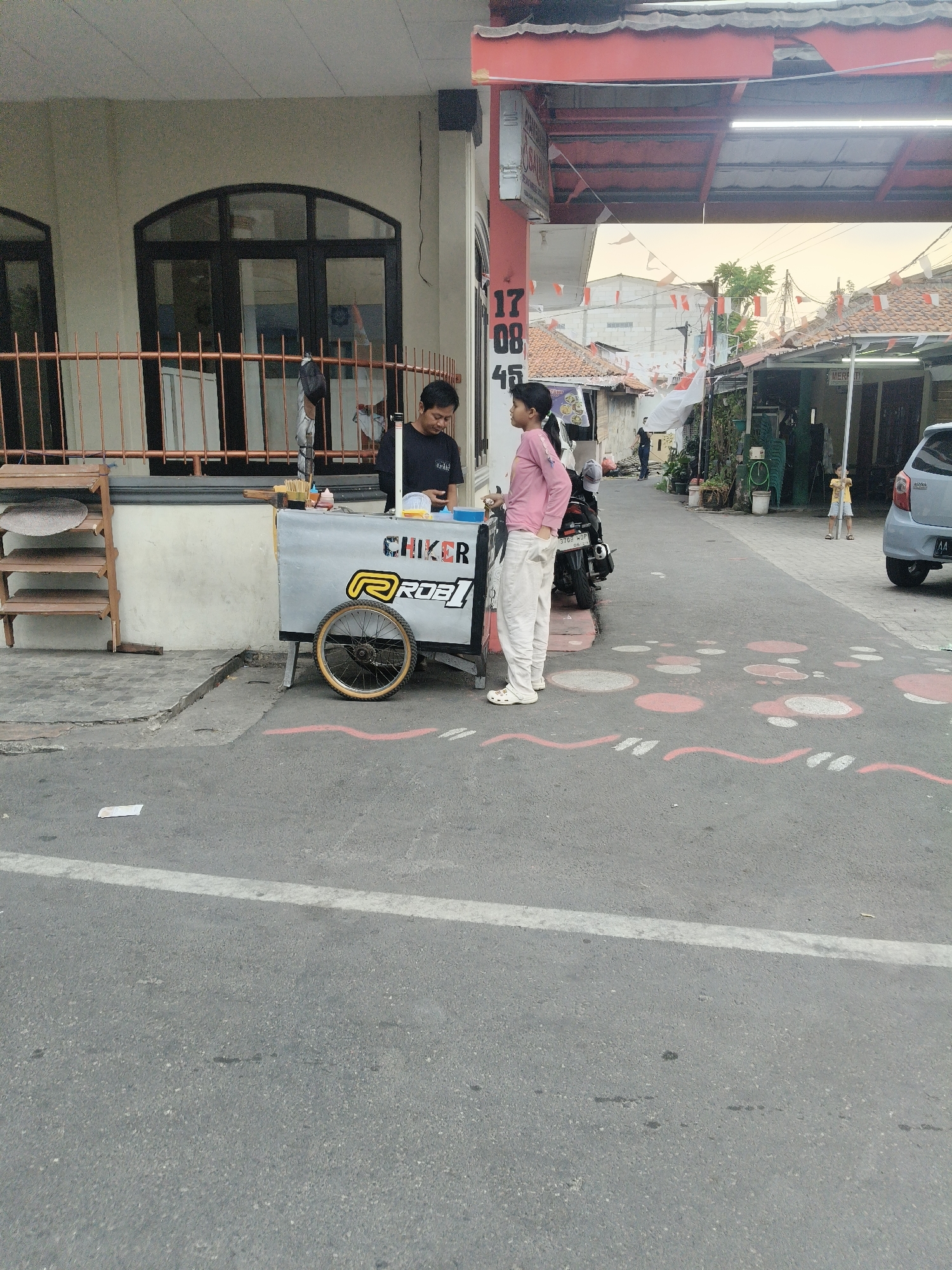 In the city of South Tangerang, Indonesia, a street scene unfolds. A man, likely a vendor, stands behind a food cart labeled "CHIKER ROBI." He is wearing a black t-shirt and appears to be engaged with a customer. A young woman in a pink long-sleeved shirt and white pants stands in front of the cart, her hands in her pockets, seemingly waiting for a purchase. The food cart is adorned with a yellow and red logo.
To the left of the cart, a wooden rack holds flat, circular items. A metal fence separates the building from the street. The building itself is a light beige color with dark-framed arched windows. In the background, houses with red tile roofs line the street, and Indonesian flags, red and white, are strung up, suggesting a local celebration or event. A motorcycle is parked behind the young woman.
Further down the street, a light blue car is visible, parked on the right side of the road. Another young person, dressed in yellow, stands near the car. The asphalt road is marked with white lines and decorative circles and wavy lines in red and white. The overall environment suggests a residential street in an Indonesian town during daylight hours, possibly in the late afternoon given the soft lighting.
