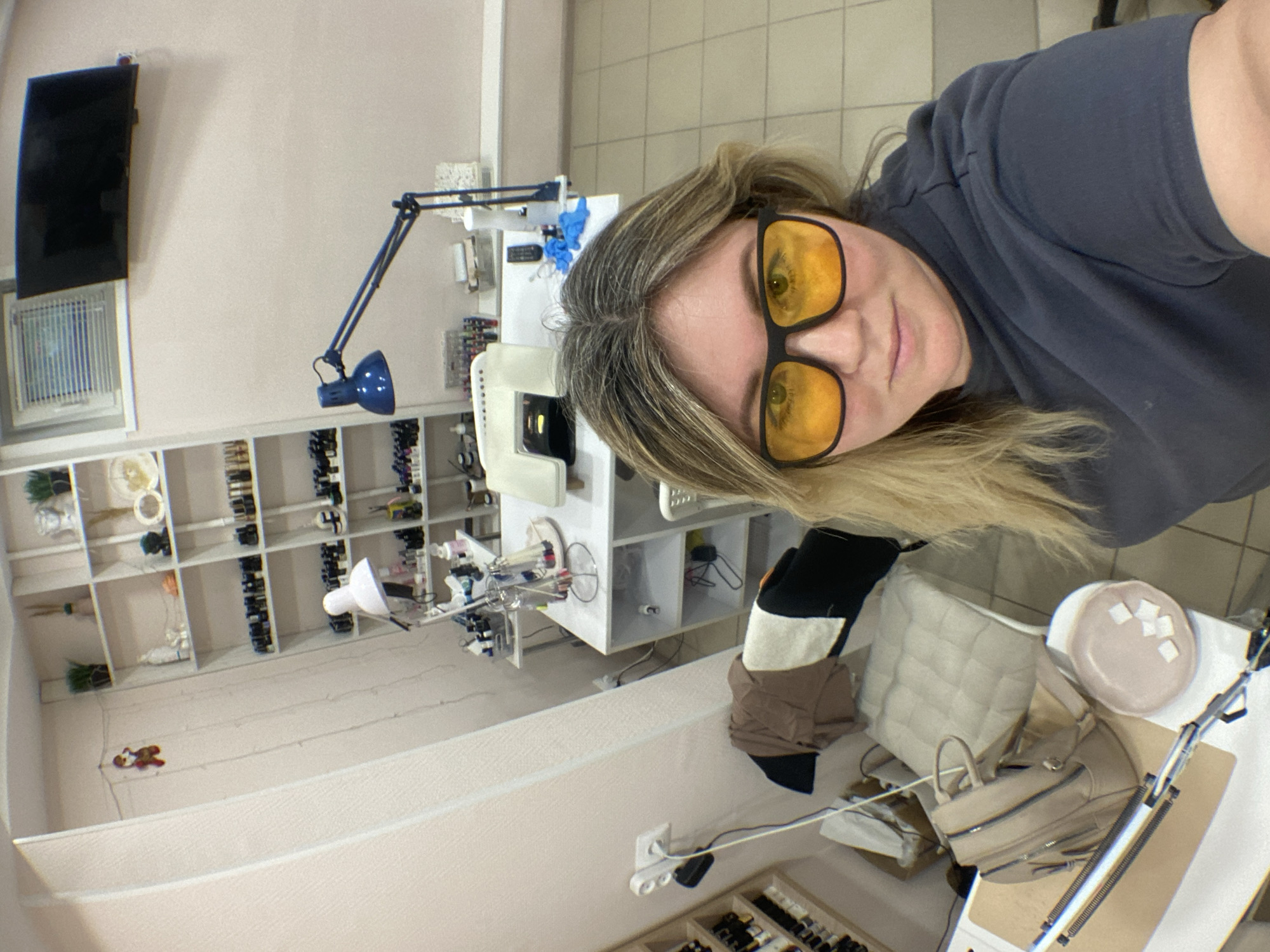 The image shows a woman wearing yellow-tinted glasses taking a selfie. She is in what appears to be a beauty salon or nail salon. Behind her, there is a shelf filled with nail polishes and other beauty products. A table with equipment and various items, including what appears to be a handbag and some padding, is also visible. A blue articulated lamp is seen mounted on the wall. Based on the available information, it is not possible to confirm the specific location in the city of Chaikovsky, Russia.