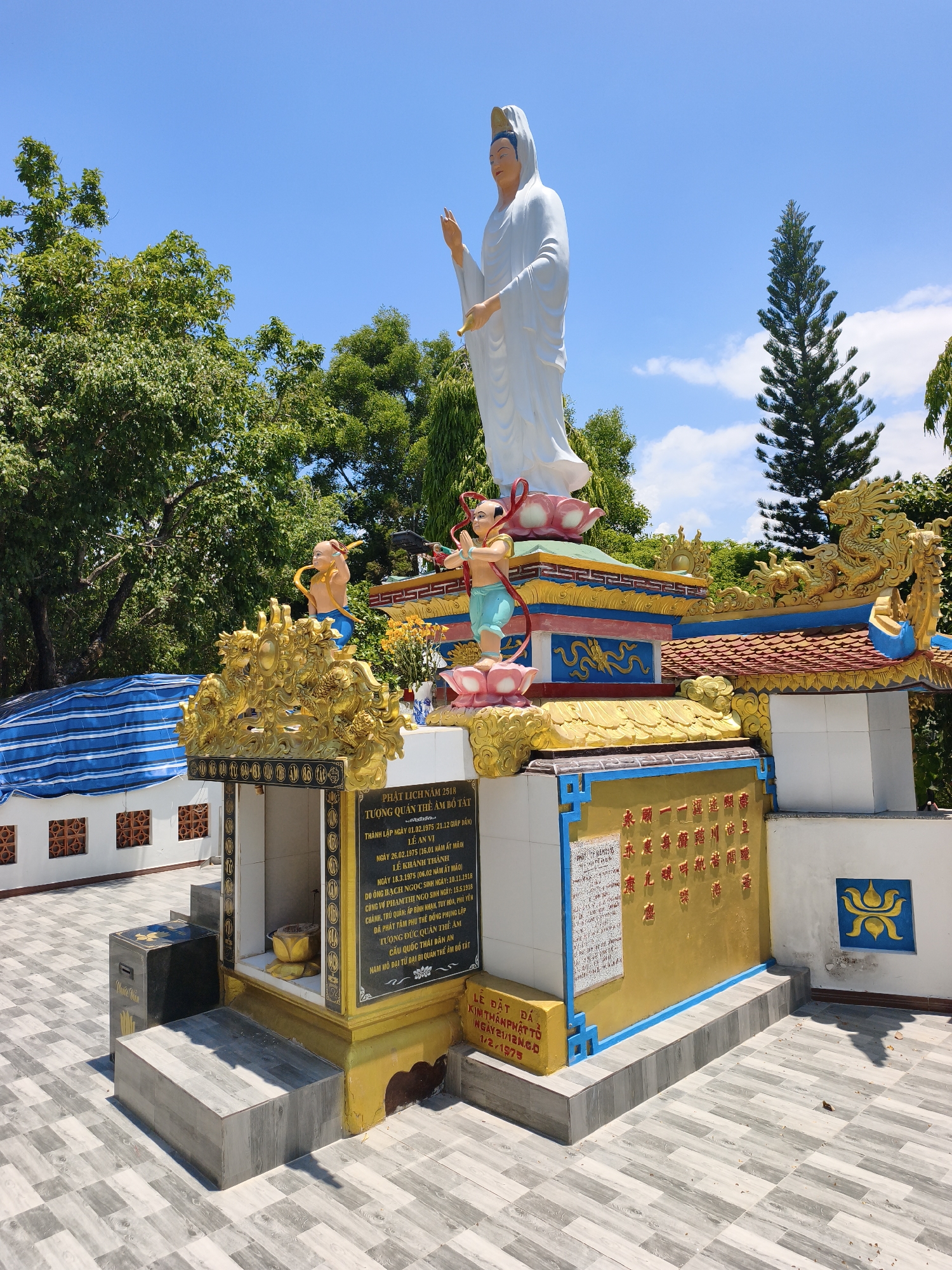 The image shows a statue of Guan Yin, the Buddhist goddess of mercy, in the city of Tuy Hoa, Vietnam. The statue is white and stands on a colorful pedestal adorned with gold dragons and other decorative elements. Two smaller figures, likely children, are positioned on either side of the main statue. Behind the statue, there are trees and a clear blue sky with some clouds. The foreground shows a tiled walkway. There are inscriptions on plaques, some in Vietnamese and some in Chinese characters, detailing historical and religious information. The overall scene appears to be part of a temple or religious site.