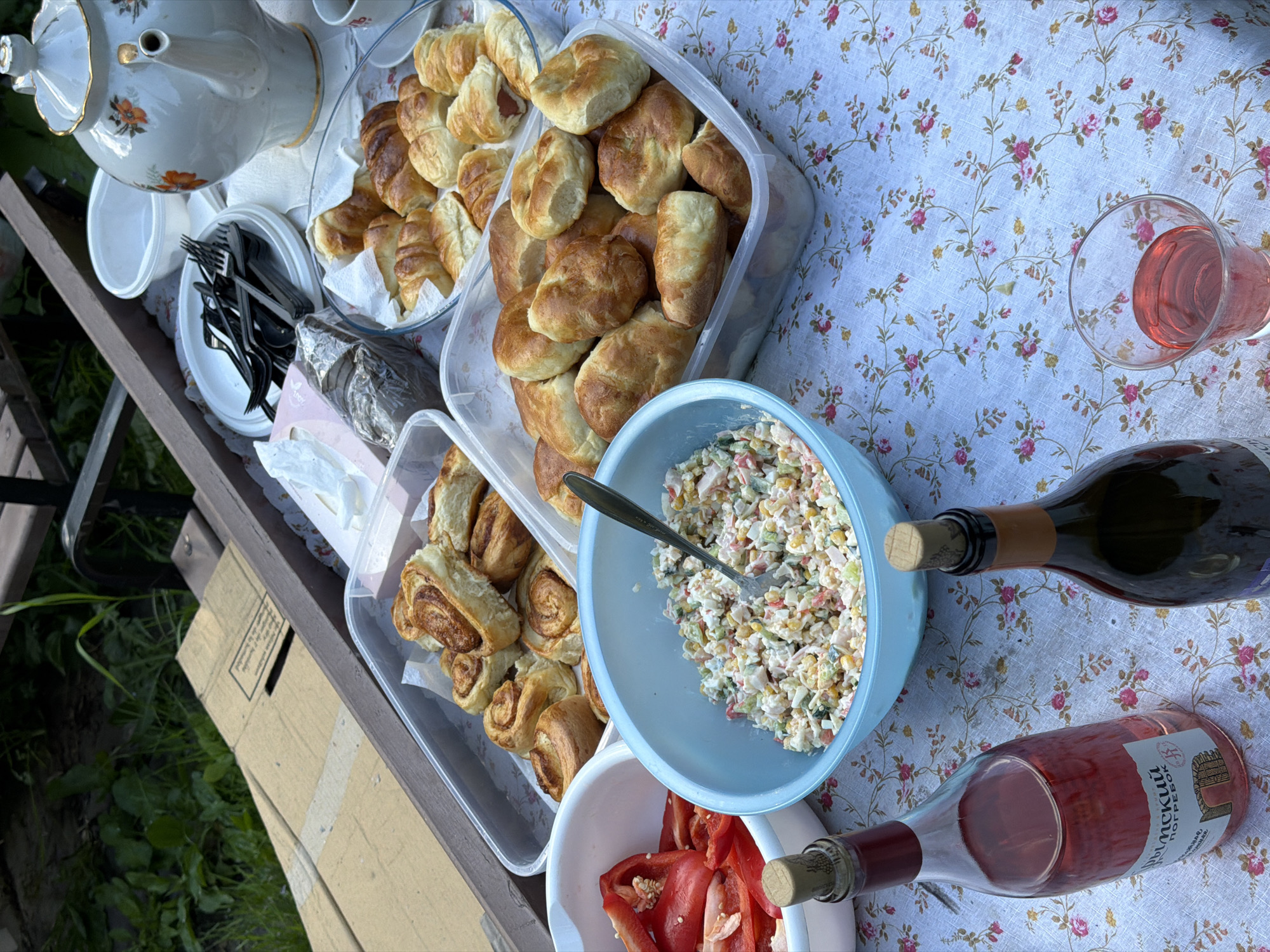 The image shows an outdoor picnic table with a floral tablecloth. On the table are various food items including pastries in clear containers, a bowl of salad with a spoon, and a bowl of sliced red peppers. Two bottles of wine and a glass of pink liquid are also present. A teapot and plates with cutlery are visible in the background. The table is on a wooden structure near a grassy area with some greenery. The label on one of the wine bottles says 'Трудовой погребок' which translates to "Trudovoy Cellar." This suggests a location related to the city of Trudovaya, Russia.