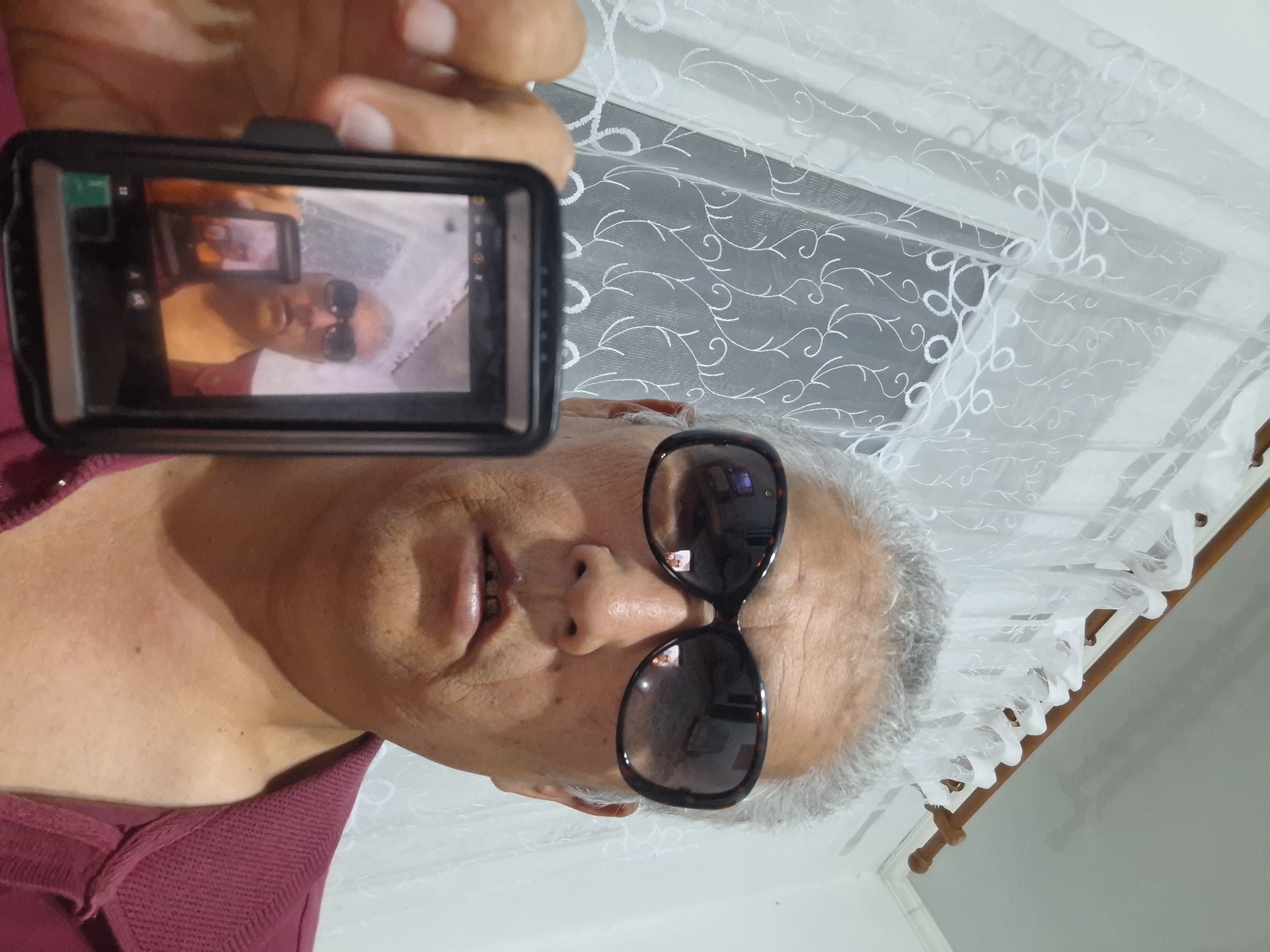 The image shows a close-up, point-of-view shot of a man holding a digital camera and taking a picture of himself, creating a recursive image. The man, who appears to be middle-aged with graying hair and wearing large, dark sunglasses and a maroon shirt, is the primary subject. He is holding a black digital camera in his left hand, and the camera's screen displays a reflection of himself, also wearing sunglasses. The background consists of a white, sheer curtain with an embroidered floral pattern, suggesting an indoor setting. The lighting is diffused, likely from natural light coming through a window behind the curtain. There are no identifiable landmarks or contextual elements to suggest a specific location within Almaty, Kazakhstan. The image captures a moment of self-portraiture using a digital device.