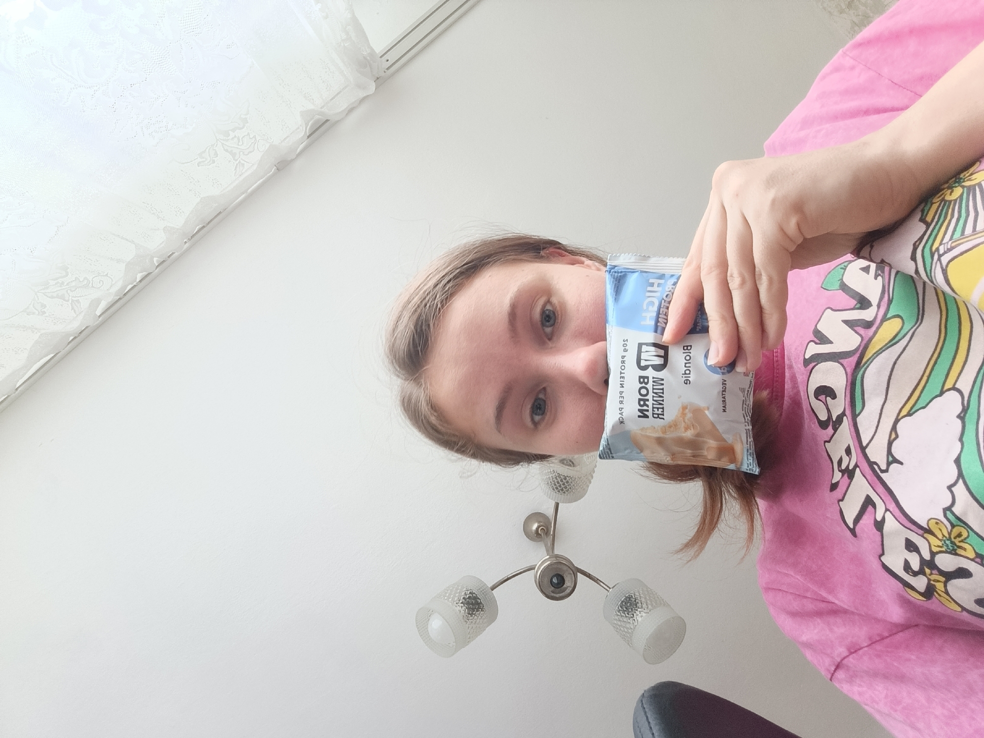 A person is holding a protein bar in front of their face, looking at the camera. The person has light brown hair and blue eyes, and is wearing a pink t-shirt with a colorful graphic design on it. The background is a white wall with a lace curtain in the upper left corner. A ceiling light fixture is visible below the person's face. The image appears to be taken indoors, possibly in a bedroom or living room. There is no information in the image to suggest it is located in Razgrad, Bulgaria.