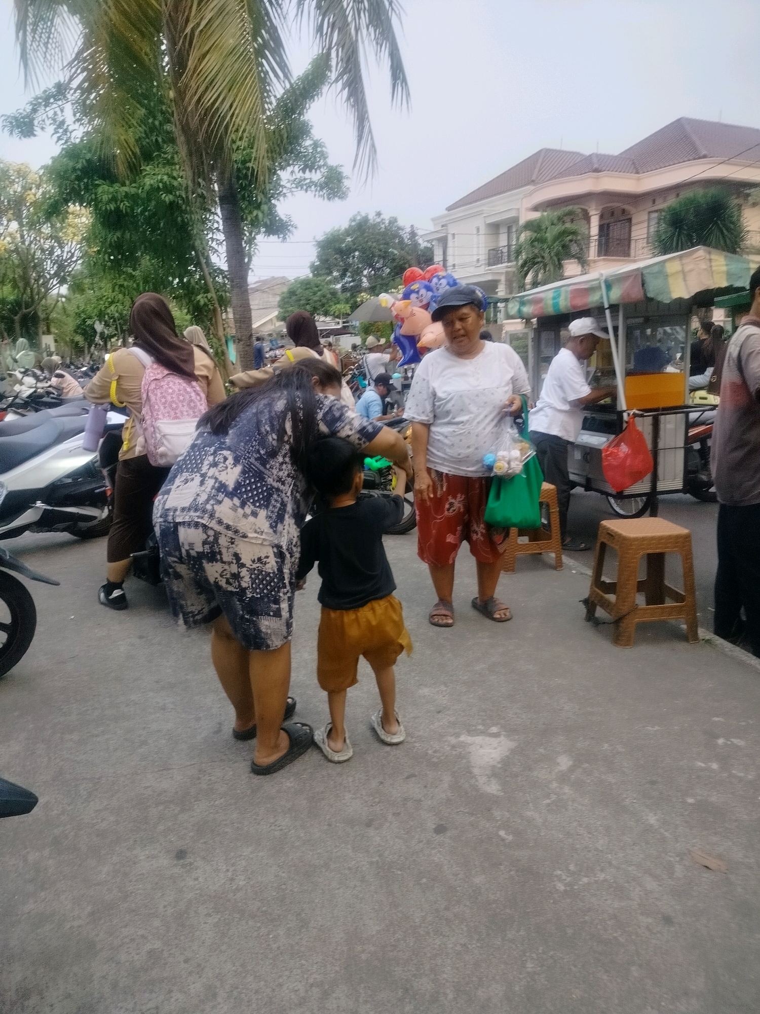 An outdoor scene in East Jakarta, Indonesia, shows a street with a group of people and parked motorcycles. In the foreground, a woman in a blue and white patterned dress is bent over, looking at a young child in an orange shorts and black t-shirt who is standing. To the left of the child, a woman in a brown hijab and beige shirt, carrying a pink backpack, is partially visible. To the right, a woman wearing a white t-shirt and red shorts stands with a green bag filled with items. Behind her, a man in a white shirt and hat is attending to a food cart, and a red plastic bag hangs from its side. Two small brown stools are placed near the food cart. In the background, buildings with tiled roofs and palm trees are visible, along with several parked motorcycles. The ground is paved, and the sky is overcast.