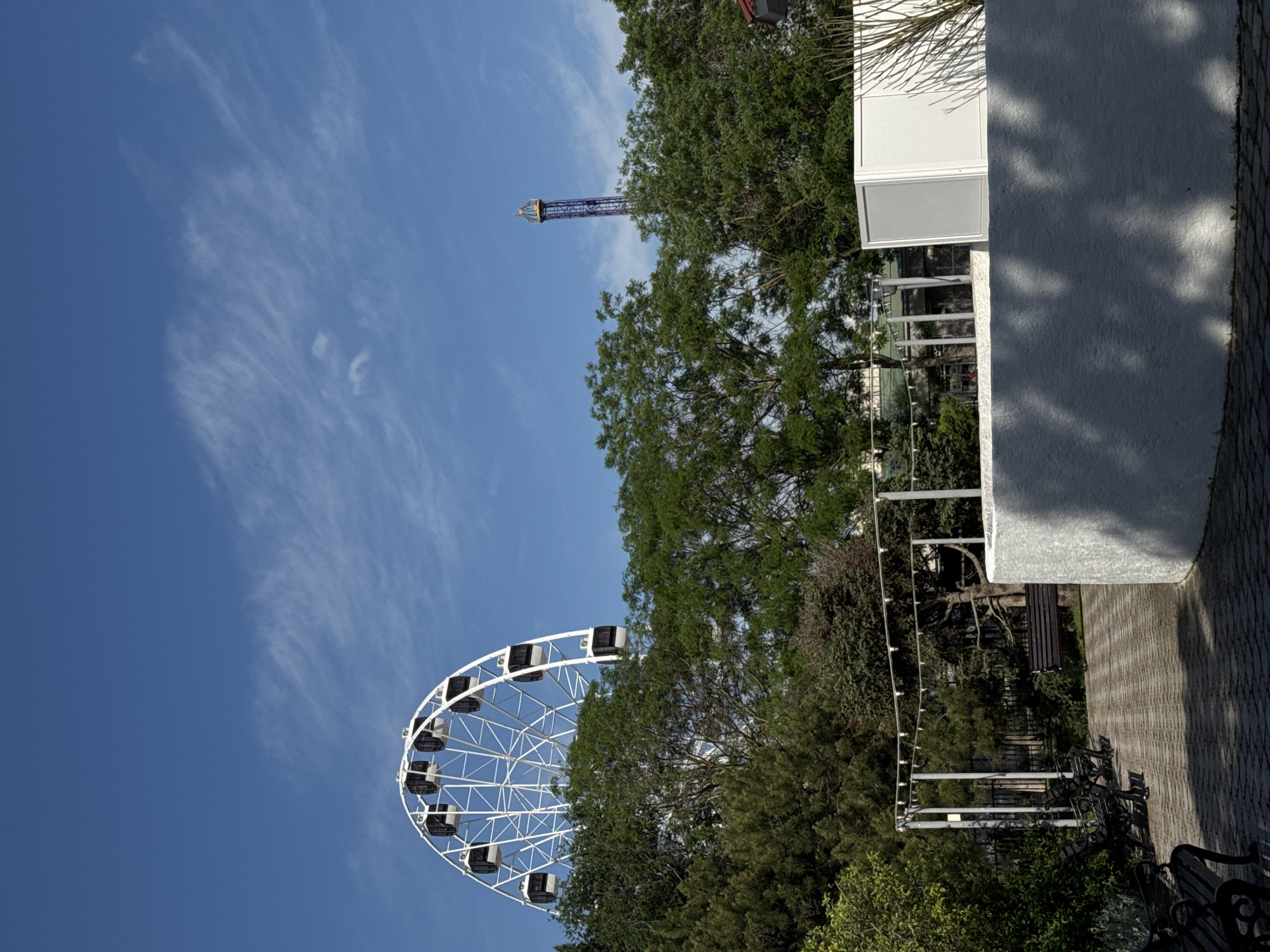 The image shows a daytime scene in what appears to be a park in Yevpatoriya, Ukraine. A white ferris wheel is visible in the lower left, set against a clear blue sky with some wispy clouds. In the center of the frame are trees, and a tall, thin tower stands in the distance. To the right, a building with white walls and a textured exterior is partially visible. A brick-paved walkway runs along the right edge, with benches and decorative lighting structures. The overall scene suggests a recreational area or amusement park setting.