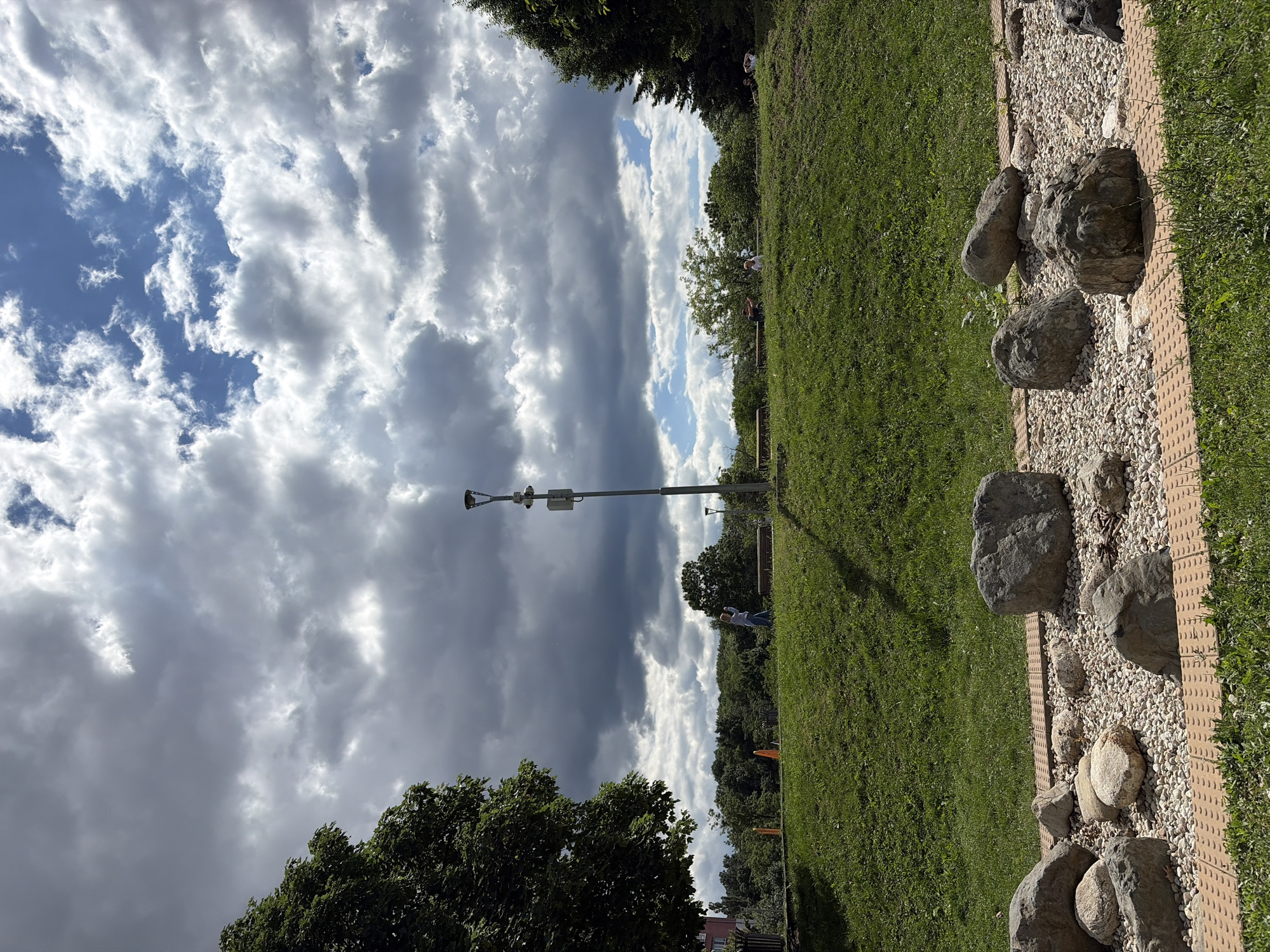 The image captures a park scene with a grassy hill under a dramatic sky. Large, gray clouds are interspersed with patches of bright blue sky, suggesting dynamic weather. In the foreground, a decorative arrangement of large, gray stones sits on a bed of smaller white and tan pebbles, bordered by a reddish-brown textured pathway. The middle ground features a gently sloping green lawn. A single lamppost with two lights and a small electrical box stands on the lawn. Further back, several people are scattered across the park. On the left, a person in a light blue top and dark pants stands near a park bench. Two more people, one adult and one child, are visible near another bench in the center. On the right side of the hill, three people are sitting or lying on the grass. A dense line of trees forms the background, with a sliver of a building visible on the far left. The overall impression is of a park on a day with changeable weather.