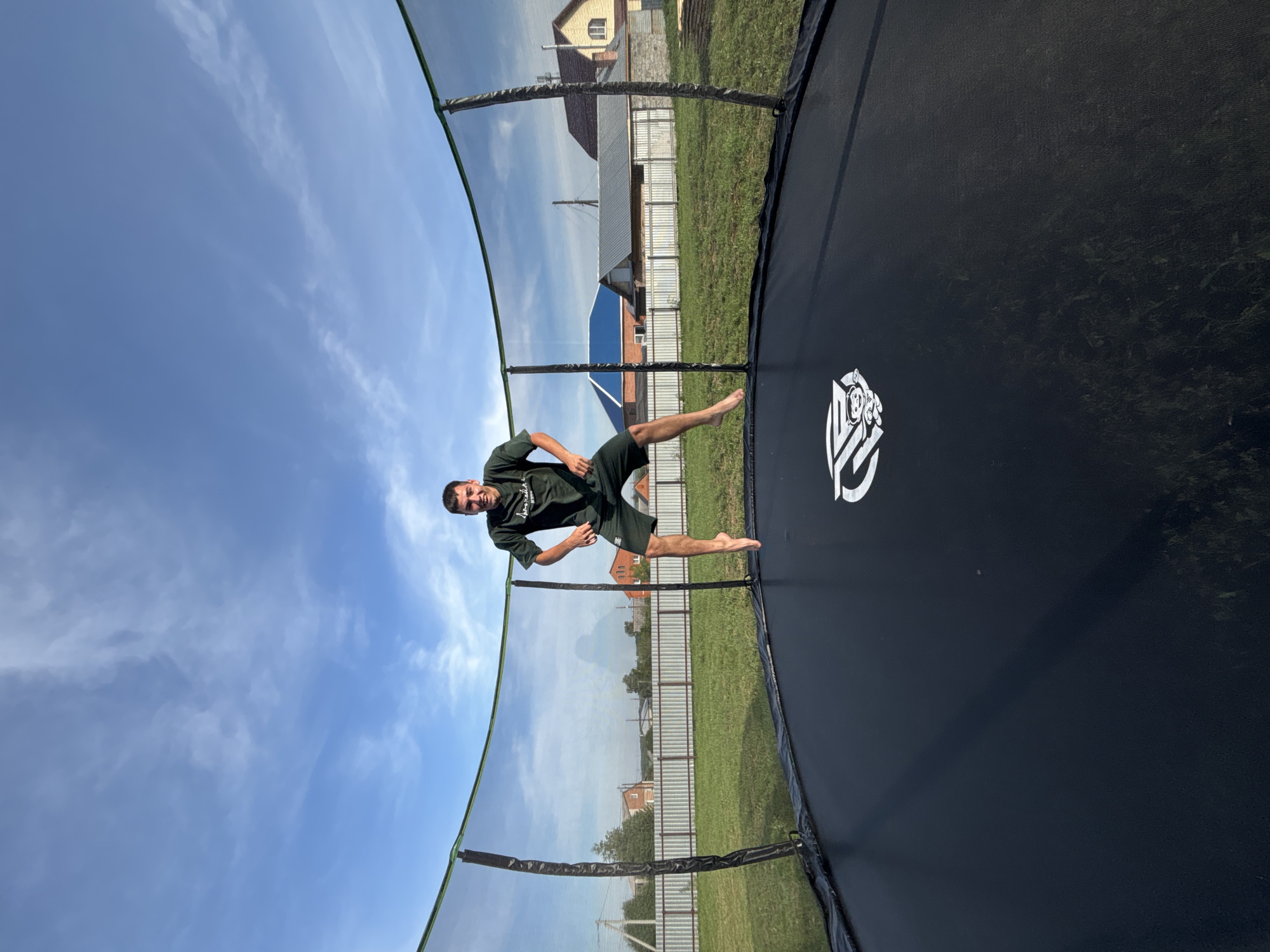 A young man is captured mid-air, jumping on a black trampoline in a backyard. He is wearing a dark green t-shirt and shorts. The trampoline is surrounded by a safety net. In the background, there is a green lawn, a white fence, and several houses with tiled roofs under a blue sky with scattered white clouds. The location is specified as Vishnevka, Russia.