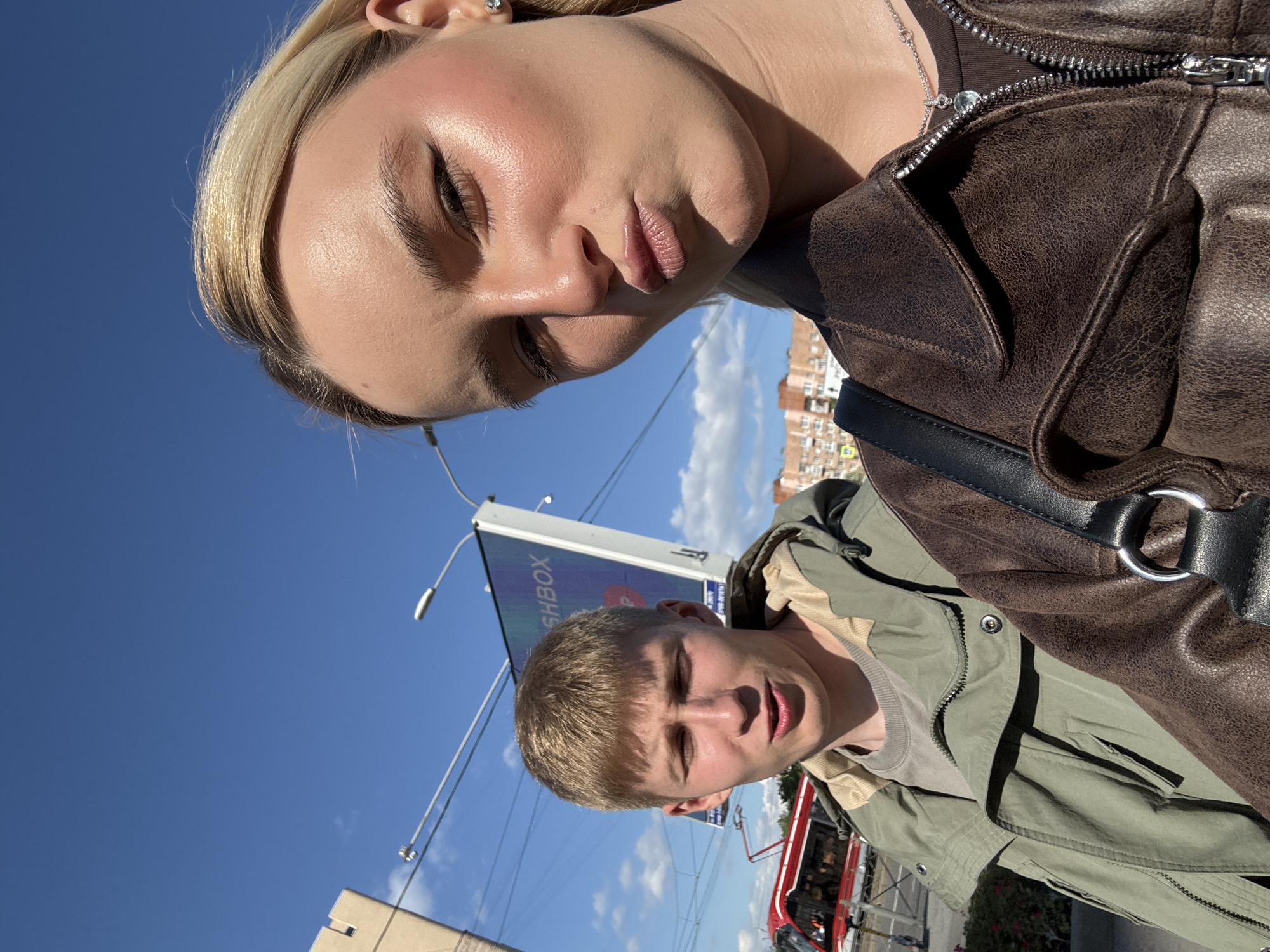 Two people are in a selfie taken outdoors. The person in the foreground is a fair-skinned woman with blonde hair wearing a brown leather jacket and a silver necklace. Her expression is a pout. Behind her is a fair-skinned man with blonde hair wearing a green jacket. His mouth is open. The sky is blue with some clouds. In the background, there is a billboard and a red and white tram, placing the location in an urban area, potentially Vsevolozhsk, Russia, given the red tram and urban characteristics. Street lights are visible in the background.