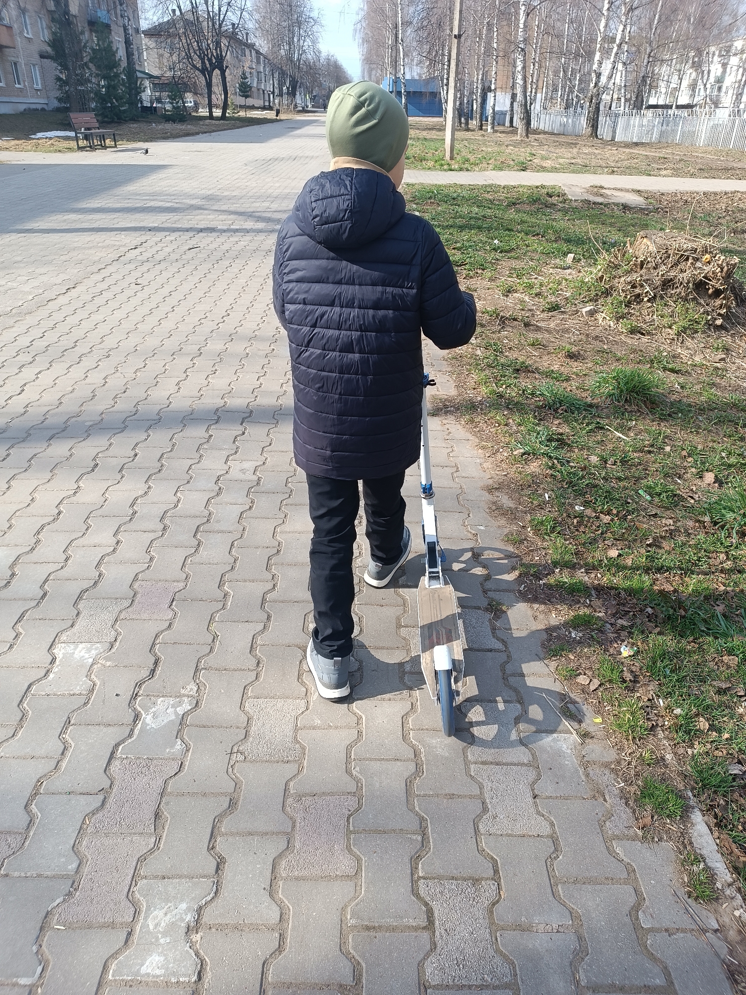 A young person is standing on a brick-paved sidewalk next to a patch of grass, holding a scooter. The person is facing away from the viewer, wearing a dark blue puffer jacket, black pants, grey shoes, and a green beanie. The scooter is mostly white with blue and light brown accents. The location is Chaikovsky, Russia. A park with bare trees and buildings are visible in the background.