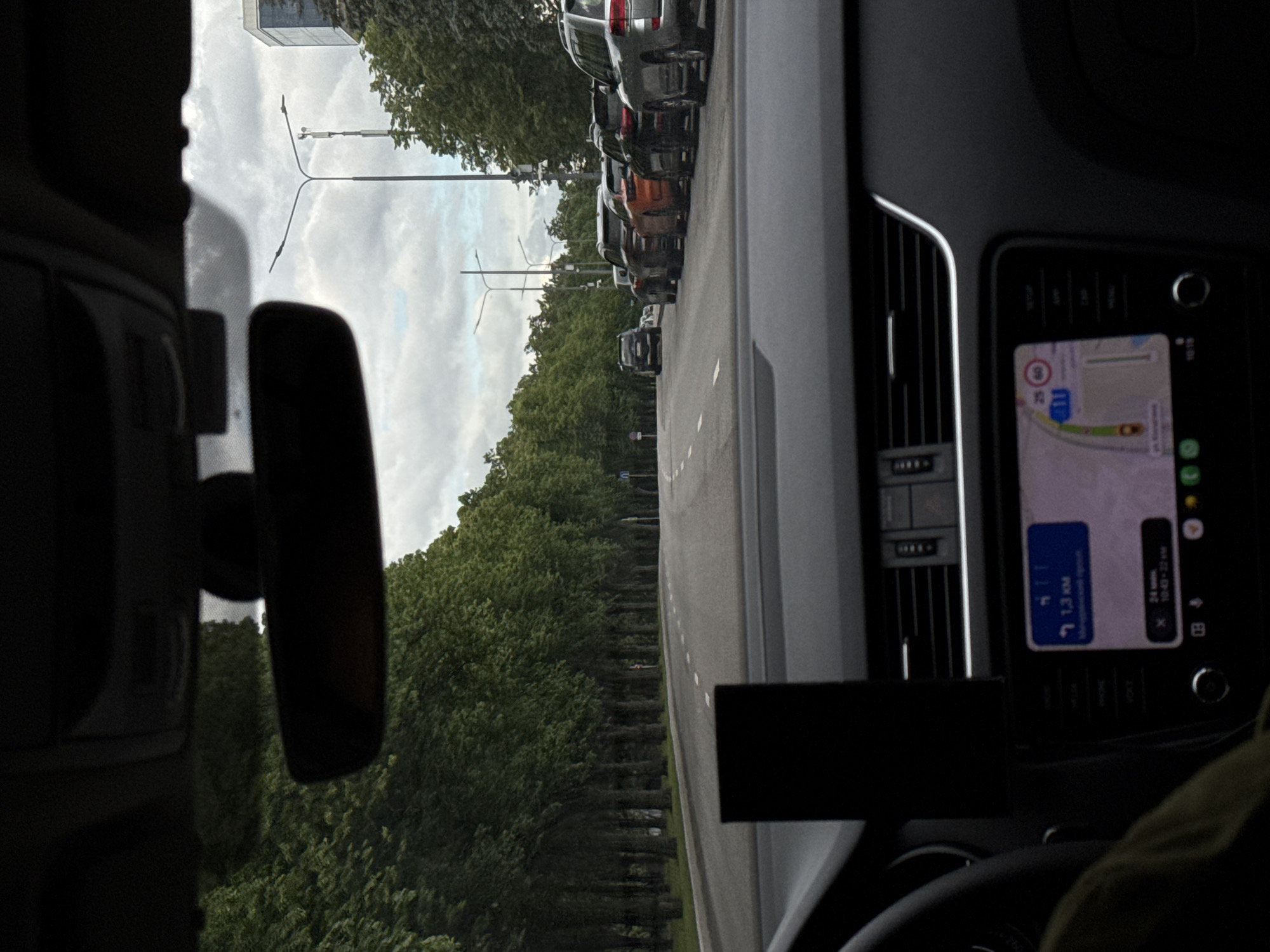 The image, taken from inside a car, shows a street scene in Moscow, Russia. The car is stopped in traffic on a road lined with dense green trees. A row of parked cars is visible to the right. The car's GPS navigation system displays a route, indicating the destination is 1.3 kilometers away. The time on the navigation system is 10:42. The sky is overcast.