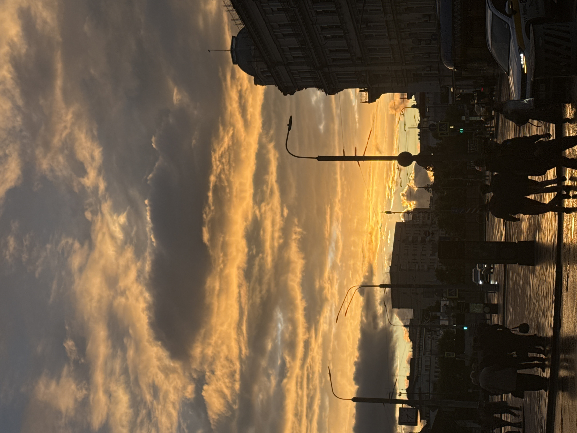 This image depicts a street scene in Moscow, Russia, likely at sunset. The sky is filled with striated clouds illuminated by golden light. Silhouetted pedestrians are visible on the sidewalk and crosswalk. The street appears wet, reflecting the ambient light. In the background, buildings are silhouetted against the bright sky, and street lamps are also present. A vehicle, possibly a taxi, is partially visible on the right side of the frame. The overall mood is one of urban twilight.