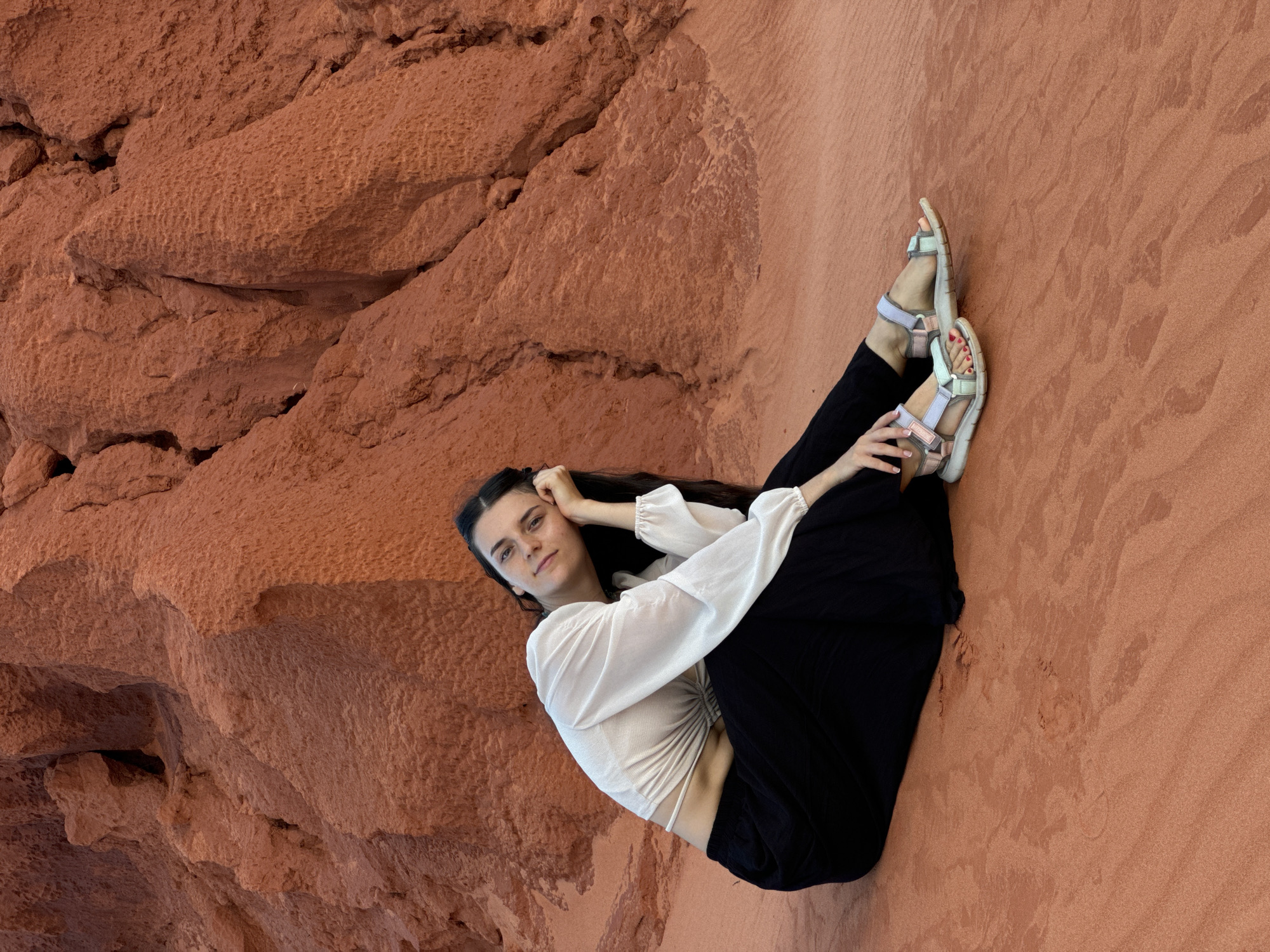 A young woman is depicted lying on her left side against a reddish-brown natural landscape, characteristic of the red sand dunes and rock formations found in Mui Ne, Vietnam. Her body is slightly curled, with her left arm propped behind her head and her right hand resting near her left ankle. She looks directly at the camera with a calm and serene expression.
She is dressed in a white long-sleeved crop top featuring gathered or ruched detailing at the front and cuffed, slightly puffed sleeves. She wears loose-fitting black pants and light-colored (possibly pastel blue, pink, and white) strap sandals, with red nail polish visible on her toes. Her dark hair flows down beside her head.
The background is dominated by the reddish-brown geological formations. The area to the left of the woman features rough, craggy rock textures, while the area to her right shows a smoother sandy surface with subtle ripple patterns, indicative of wind-blown sand. The overall lighting is bright and even, suggesting a clear daytime setting. There is no visible text in the image.