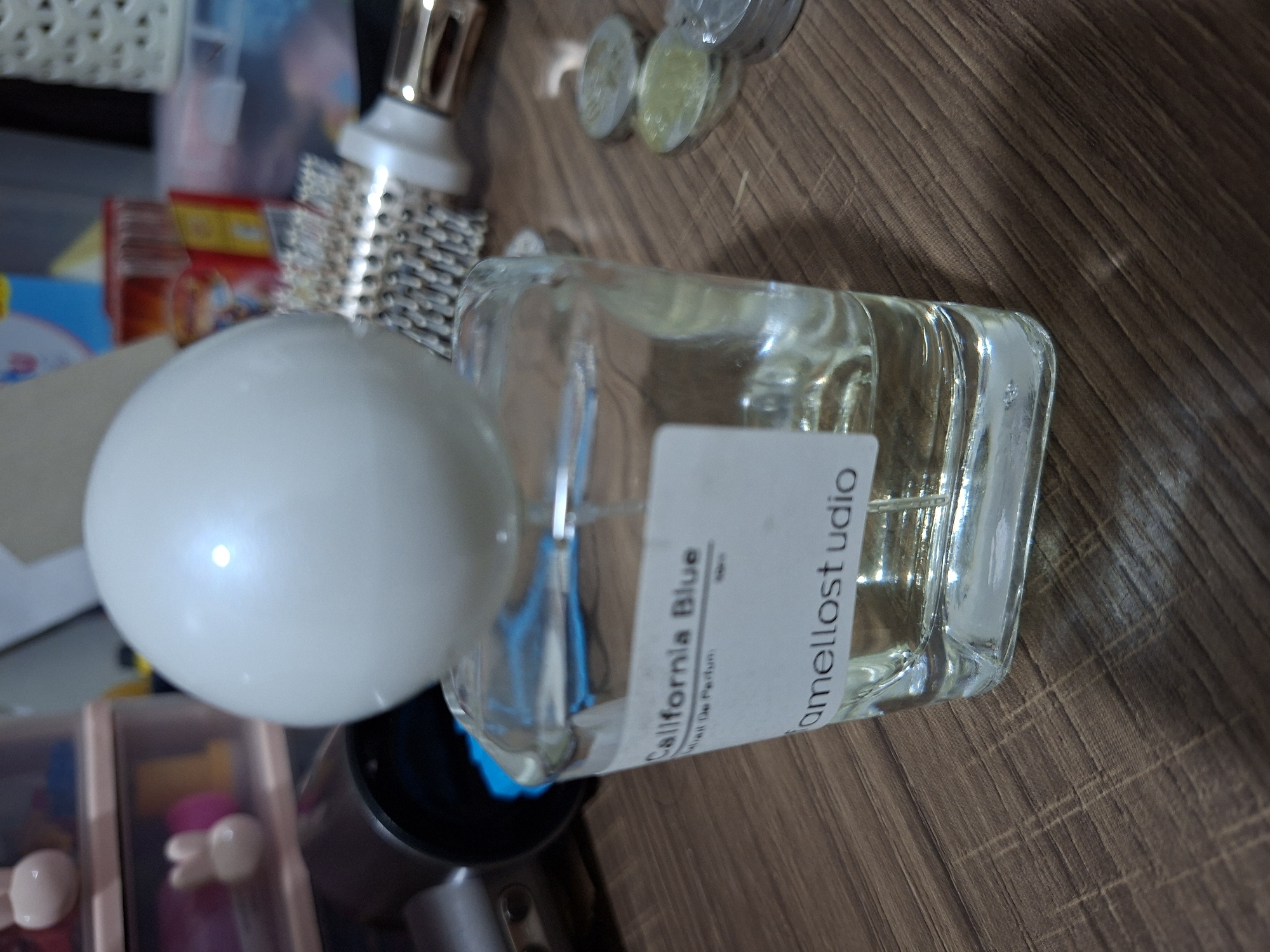 The image displays a clear glass perfume bottle with a white spherical cap, placed on a wooden surface. A white label on the bottle reads "California Blue" and "mellostudio." In the background, various objects are visible, including what appears to be a hair dryer, a hair brush, small plastic drawers with bunny-shaped knobs, and stacks of colorful boxes. Coins are scattered on the wooden surface to the right of the perfume bottle. The overall scene suggests a personal vanity or dresser area. Without further information or context, it is not possible to determine the specific location within the city of Samarinda, Indonesia.