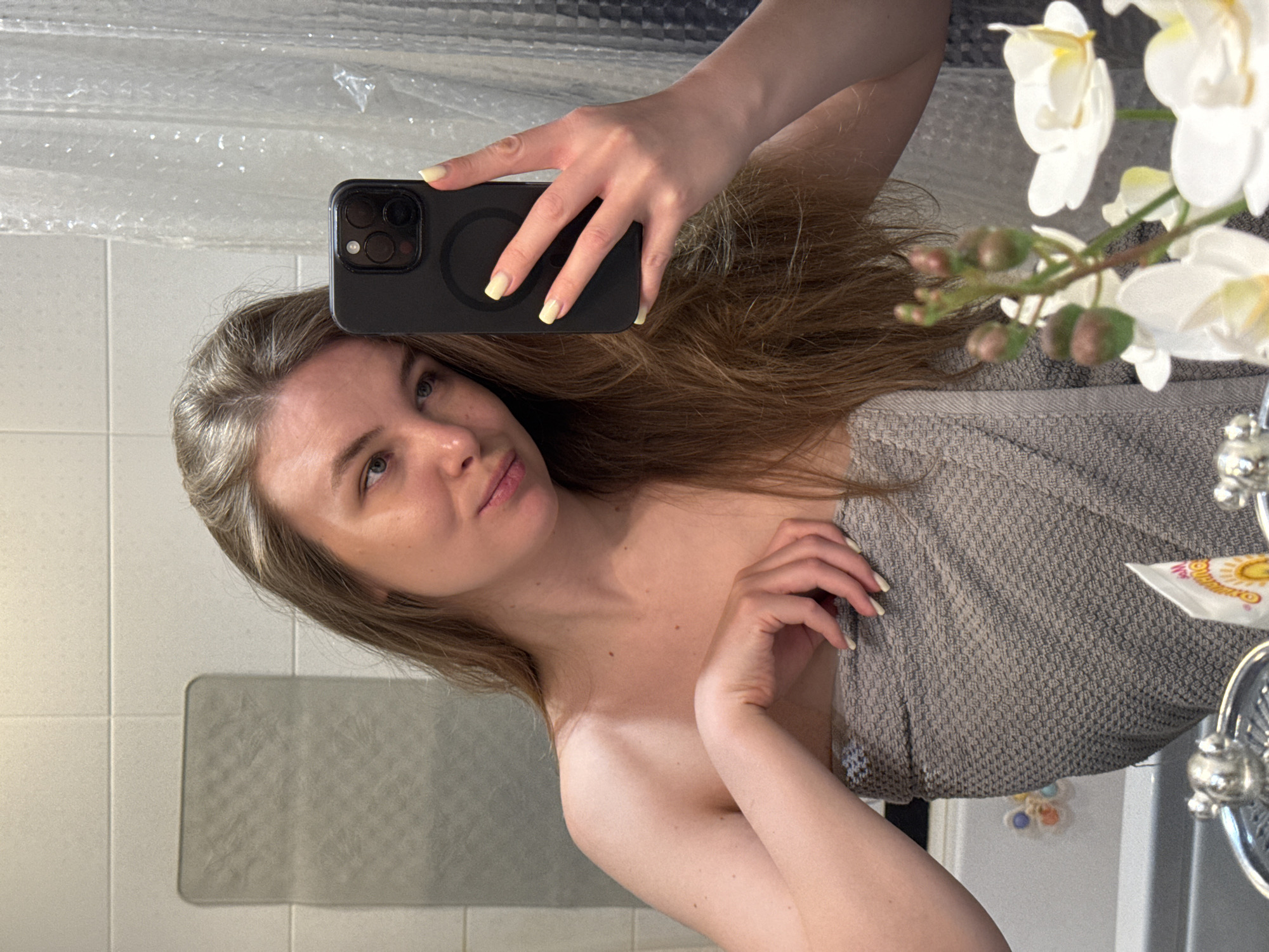 A person is taking a selfie in a bathroom. The person is partially wrapped in a grey towel and is holding a black smartphone with yellow nail polish visible. The bathroom has white tiled walls and a shower curtain. An orchid is also visible in the scene. There is no information suggesting this image was taken in Vladimir, Russia.