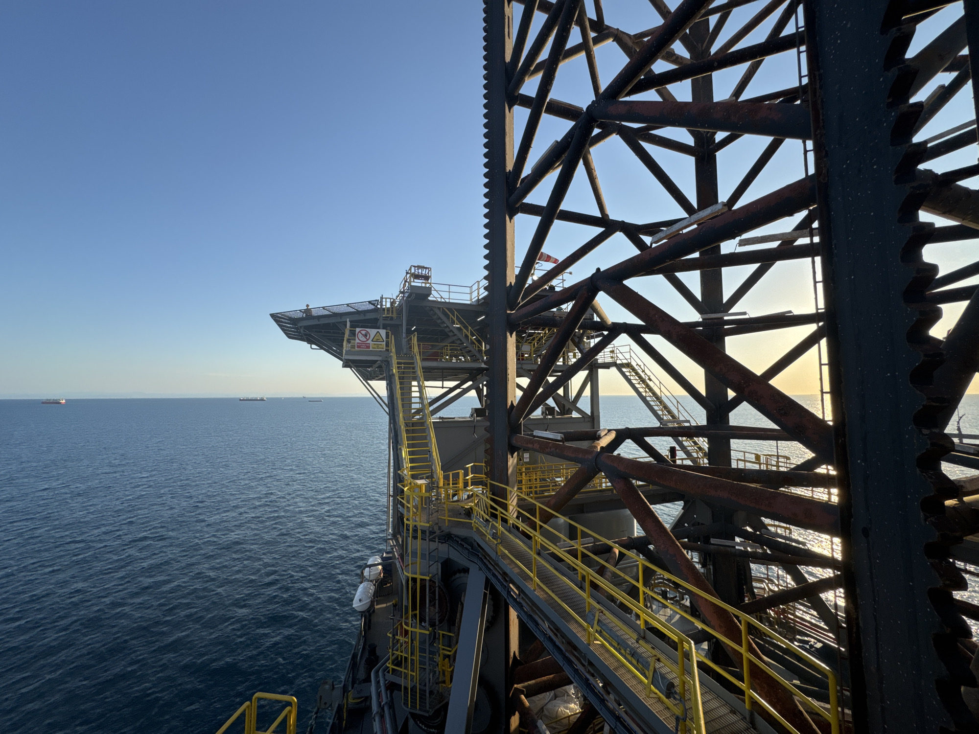 An elevated view captures a section of a large offshore structure against a clear blue sky and a calm blue sea. The structure, made of dark metal beams, features yellow railings and staircases that lead to a helideck. In the distance, several cargo ships are visible on the horizon. The sunlight glints off the water, creating bright reflections. The image is taken from a high vantage point on the structure itself, looking outwards.