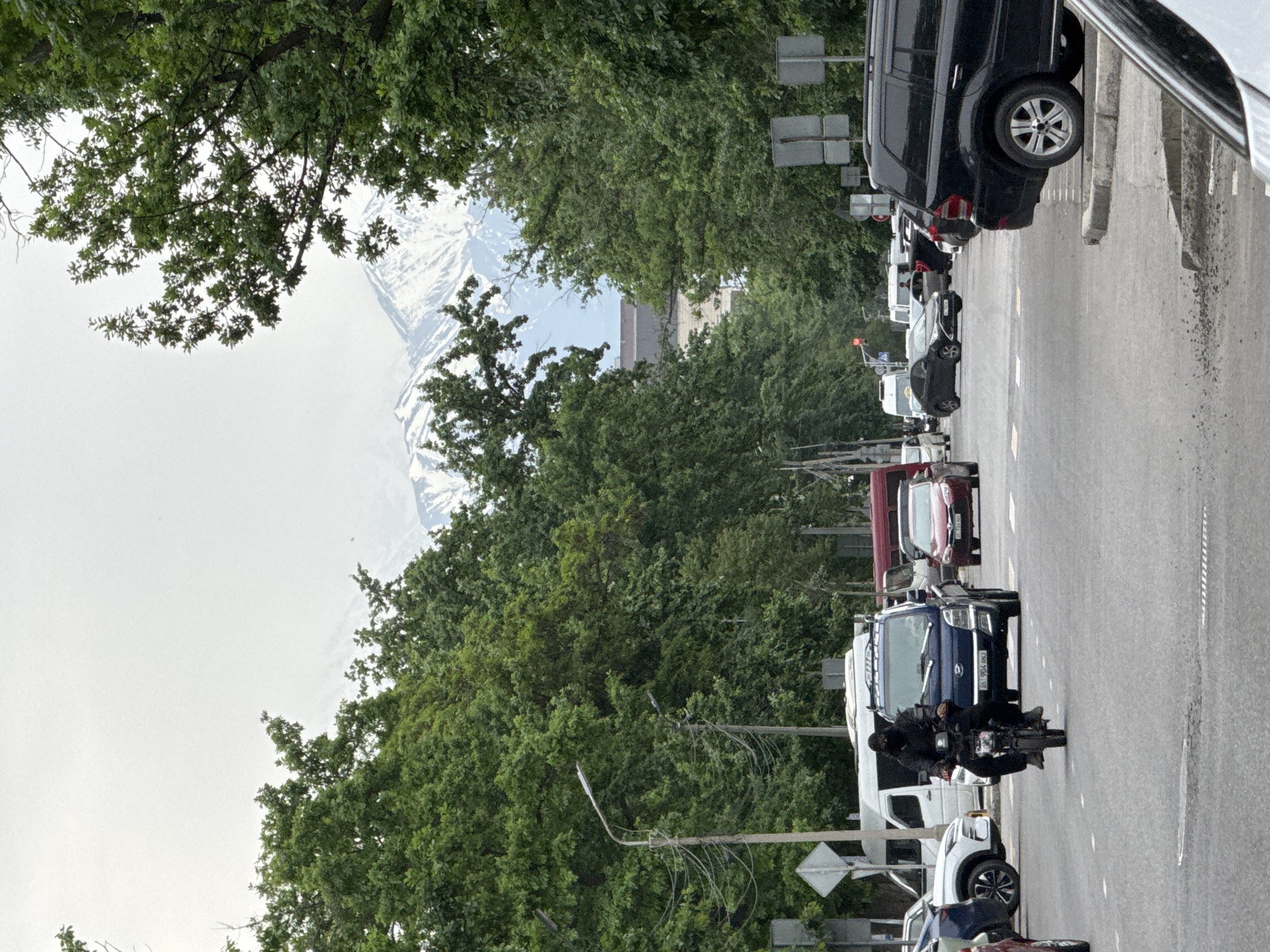 The image captures a street scene in Bishkek City, Kyrgyzstan, characterized by a line of parked cars along the right side of the road. A motorcyclist, dressed in black, is positioned on the road among the parked vehicles. The background features dense green trees partially obscuring snow-capped mountains in the distance, contrasting with the overcast sky. The street is paved and appears to be an urban thoroughfare with lampposts. The general environment suggests a moderately busy city setting with a mountainous backdrop.