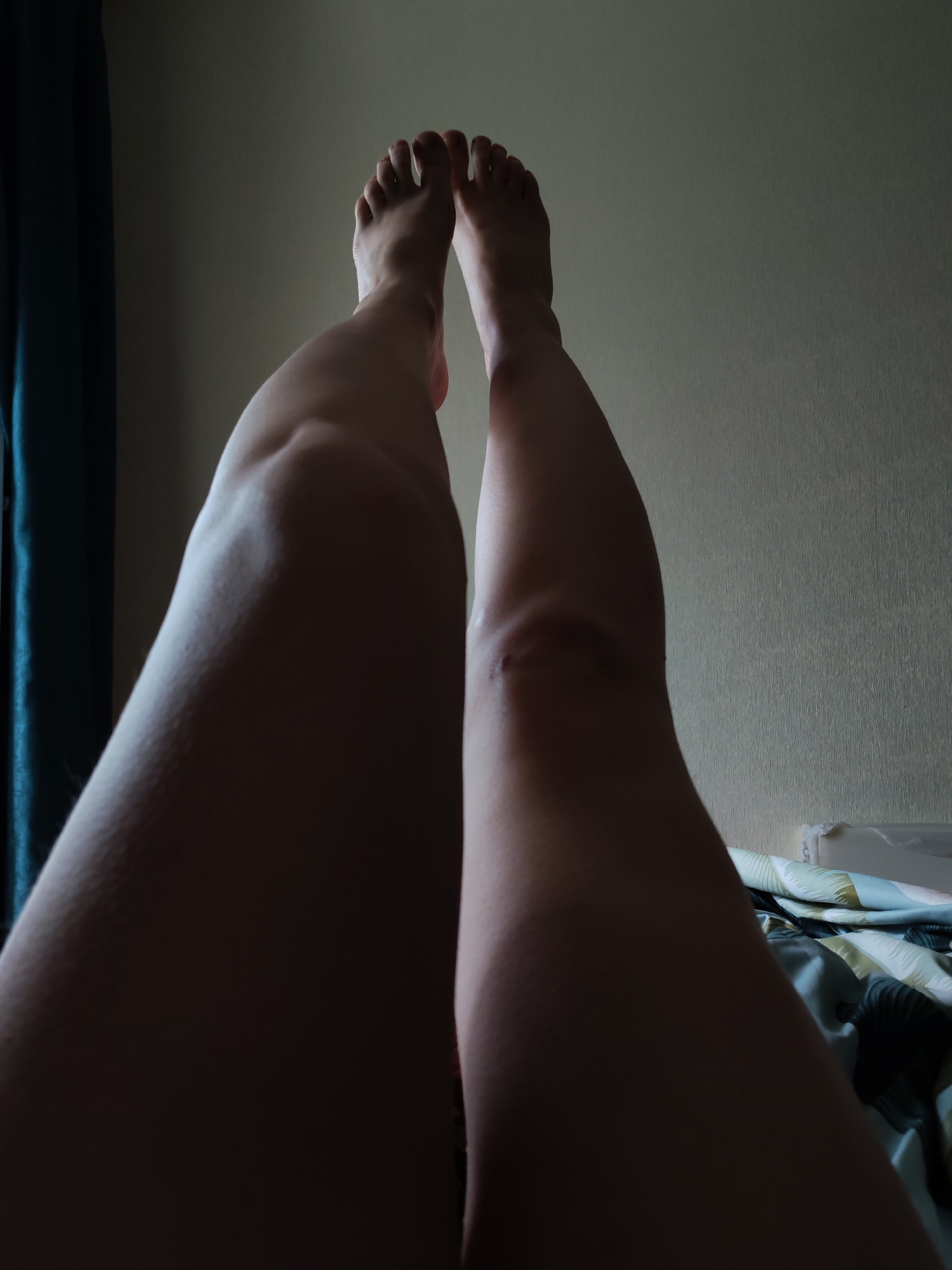 The image shows a pair of legs raised upwards, with feet visible at the top of the frame. The legs are positioned against a light-colored wall. To the left, a partially visible blue curtain hangs. Below the legs, a bed with a patterned duvet is visible. The lighting is dim, casting shadows on the legs. There is no indication of the location being Izhevsk, Russia.