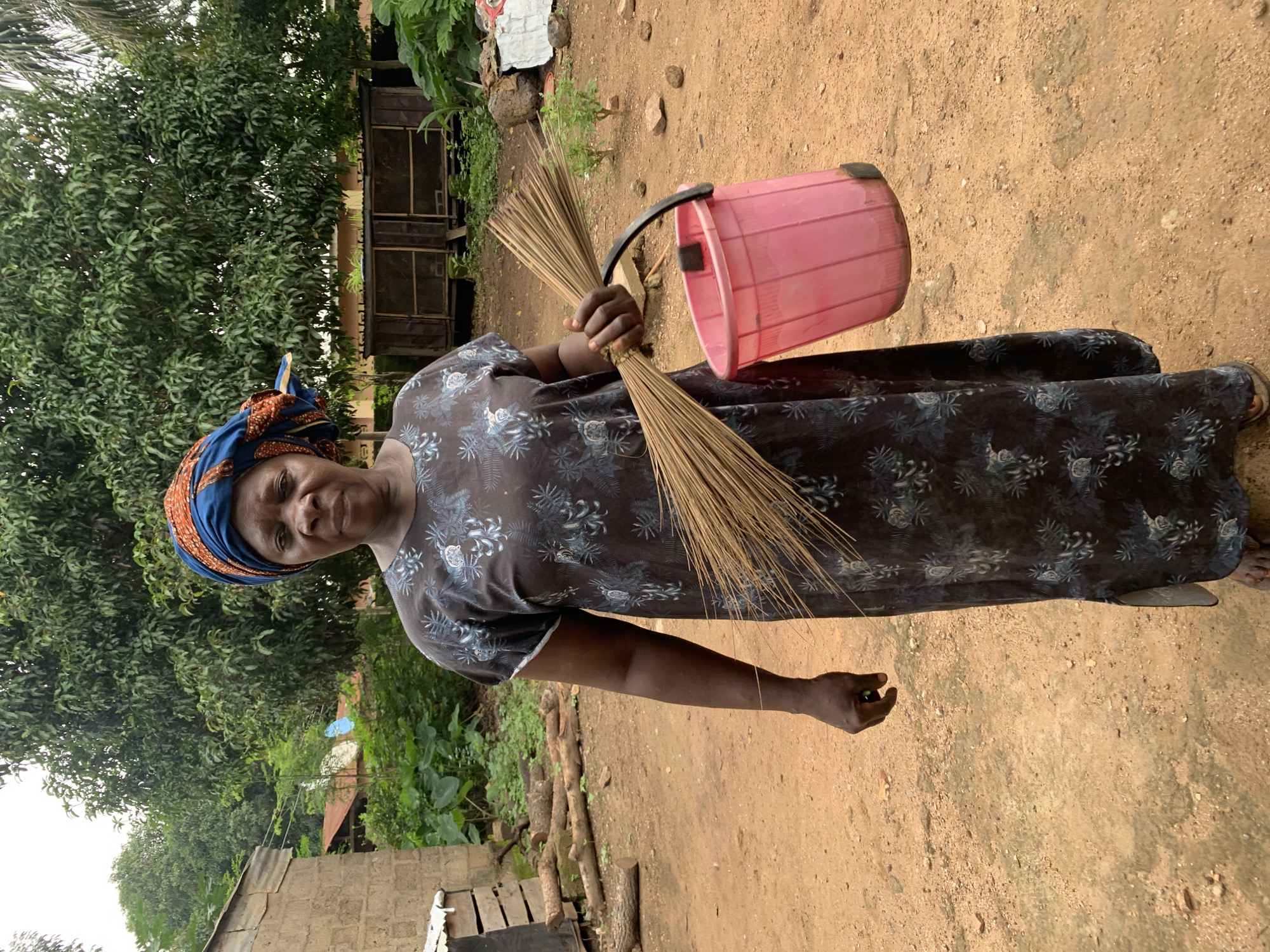 A woman stands outdoors in a dusty, rural environment, holding a broom and a pink bucket. She wears a dark, patterned dress and an orange and blue headwrap. Behind her, trees and simple structures suggest a village setting. The ground is dirt, with scattered rocks and some vegetation. The overall impression is one of everyday life in Akwanga, Nigeria.