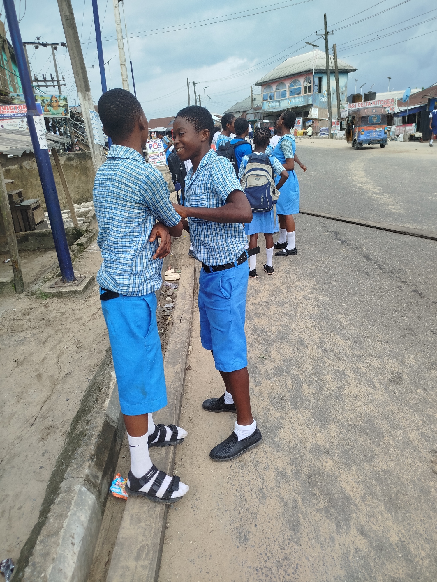 In Yenegoa, Nigeria, two young boys in matching blue and white plaid shirts and blue shorts stand on a dusty roadside, engaged in conversation. They appear to be schoolchildren, as evidenced by their uniforms and backpacks, some of which are visible on other children in the background. One boy faces the camera, smiling broadly, while the other stands slightly turned away, also seemingly looking at his friend. The scene is set on a paved road with a concrete gutter running alongside it. In the background, various buildings line the street, including one with a sign that reads "OD&J DELTA KITCHEN." A colorful three-wheeled vehicle, likely a Keke Napep, is parked further down the road. Overhead power lines crisscross the sky, suggesting an urban or semi-urban setting. The sky is overcast, indicating a cloudy day, possibly humid, which is typical for the region. The overall atmosphere appears casual and everyday, capturing a moment between two young friends on their way to or from school. Visible text on signs includes "VICTOR INTERNATIONAL SCH," "POS," and "Starines." A snack wrapper is visible on the gutter.
