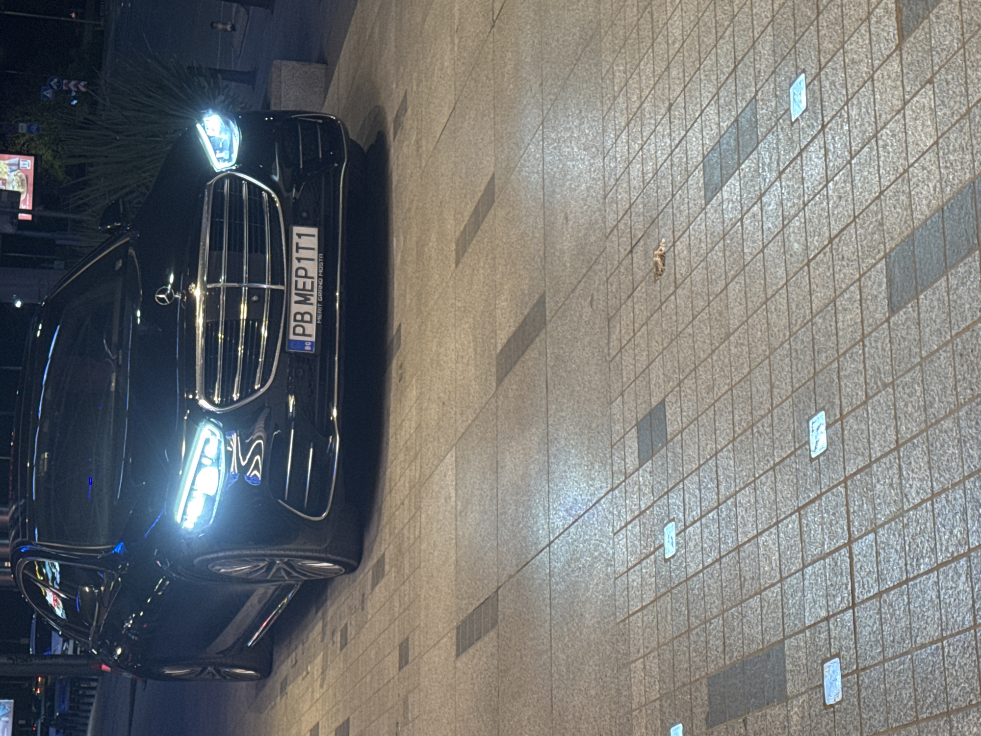 A black Mercedes-Benz with Bulgarian license plate "PB MEP1T1" is parked on a tiled pavement. The scene is outdoors, likely at night given the darkness and headlights of the car. The location is Sofia, Bulgaria, as indicated by the Bulgarian license plate. The pavement is made of square tiles with occasional rectangular gray tile inserts. There is a small plant visible in the upper left of the frame. A sign can be seen blurry in the background.