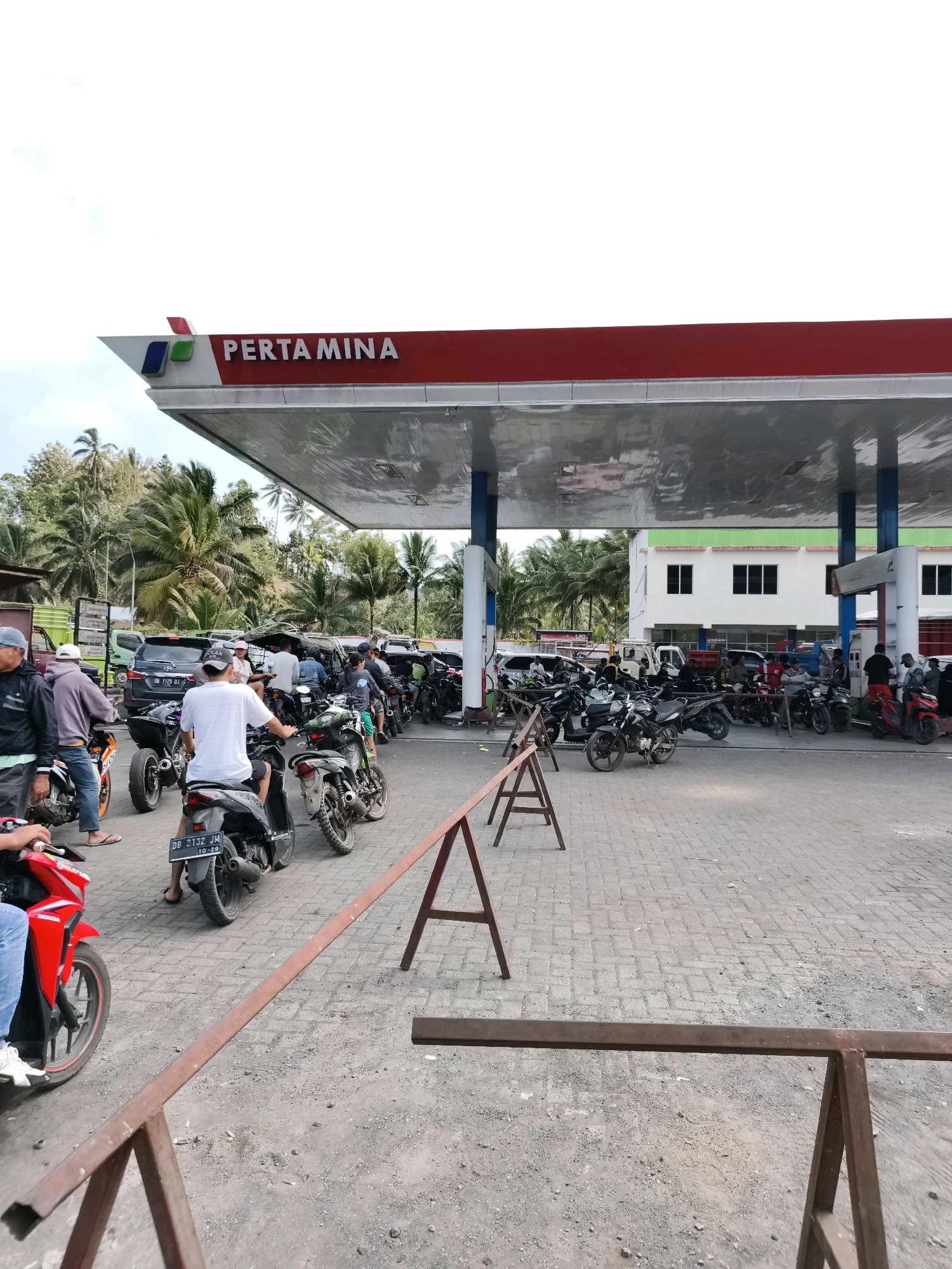 The image depicts a busy Pertamina gas station in Mundung, Indonesia, with a long line of motorcycles and a few cars waiting to refuel. The gas station has a red and white canopy with the Pertamina logo prominently displayed. Several people are seen on and around their motorcycles, indicating a normal day at the station. The environment appears to be a tropical setting, with palm trees visible in the background. The ground is paved with interlocking bricks, and there are temporary barriers set up, possibly to manage traffic flow. The sky is partly cloudy, suggesting fair weather conditions.