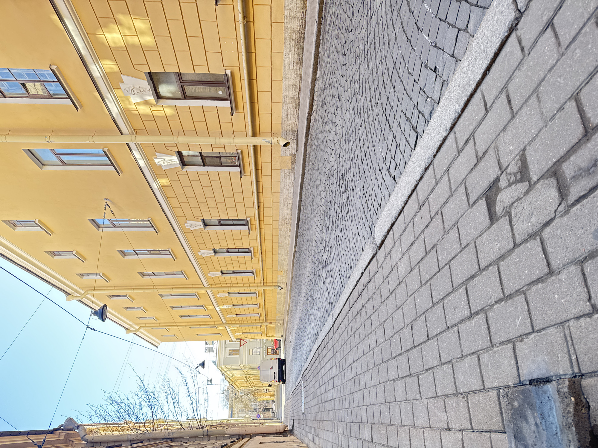 The image captures a sunny street scene in an urban environment, likely a narrow street or alleyway. The perspective is from ground level, looking down the street.
To the left, a prominent multi-story building with a light yellow facade dominates the view. It features numerous rectangular windows, some with small white decorative architectural elements or reliefs above them. Vertical downspouts are visible along the building's exterior. Sunlight strongly illuminates the building, creating distinct shadows and highlighting a tiled pattern on its upper section or roofline.
The street itself is paved with dark grey, somewhat irregular cobblestones. Adjacent to the street, on the right side of the frame, is a wide sidewalk made of lighter grey, rectangular paving stones laid in a uniform pattern. A concrete curb separates the street from the sidewalk.
Above the buildings, a clear, bright blue sky is visible, indicating good weather. Overhead wires crisscross the sky, and a single, dark-colored street lamp is suspended by these wires. In the distance, further down the street, other buildings with similar architectural styles are discernible, along with a white van and a red vehicle parked or moving. Bare tree branches are visible in the lower left foreground, suggesting late winter or early spring.
The time of day appears to be daytime, with strong sunlight creating clear shadows. On a sign visible on a distant building, the text "HARRY WANG" can be partially read. The architectural style and streetscape are characteristic of a historic European city, consistent with Saint Petersburg, Russia.