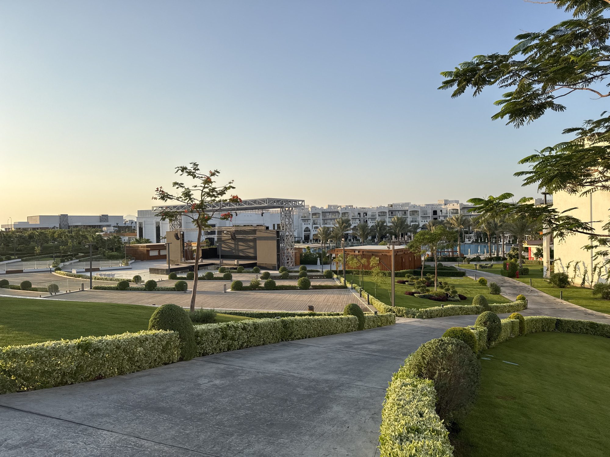 The image depicts a well-maintained resort or hotel complex in Sharm El Sheikh, Egypt. The scene is bathed in the warm, soft light of either sunrise or sunset, casting long shadows across the paved pathways and manicured green lawns. In the foreground, a curving concrete pathway leads the eye towards a central plaza area. This plaza features a stage or performance area with a modern, industrial-style metal structure. Surrounding the plaza are meticulously trimmed hedges and spherical topiaries, creating a sense of order and elegance.
In the mid-ground, a series of white, multi-story buildings with balconies line the landscape, typical of resort architecture. Palm trees are interspersed throughout the grounds, adding to the tropical ambiance. Further in the distance, more buildings and structures are visible, along with hints of a swimming pool area with lounge chairs. The sky is a clear, pale blue, transitioning to a soft yellow hue near the horizon, indicative of the golden hour. There are no people visible in the image, suggesting an unoccupied or quiet period at the resort. The overall impression is one of a serene and luxurious outdoor space, designed for relaxation and entertainment.