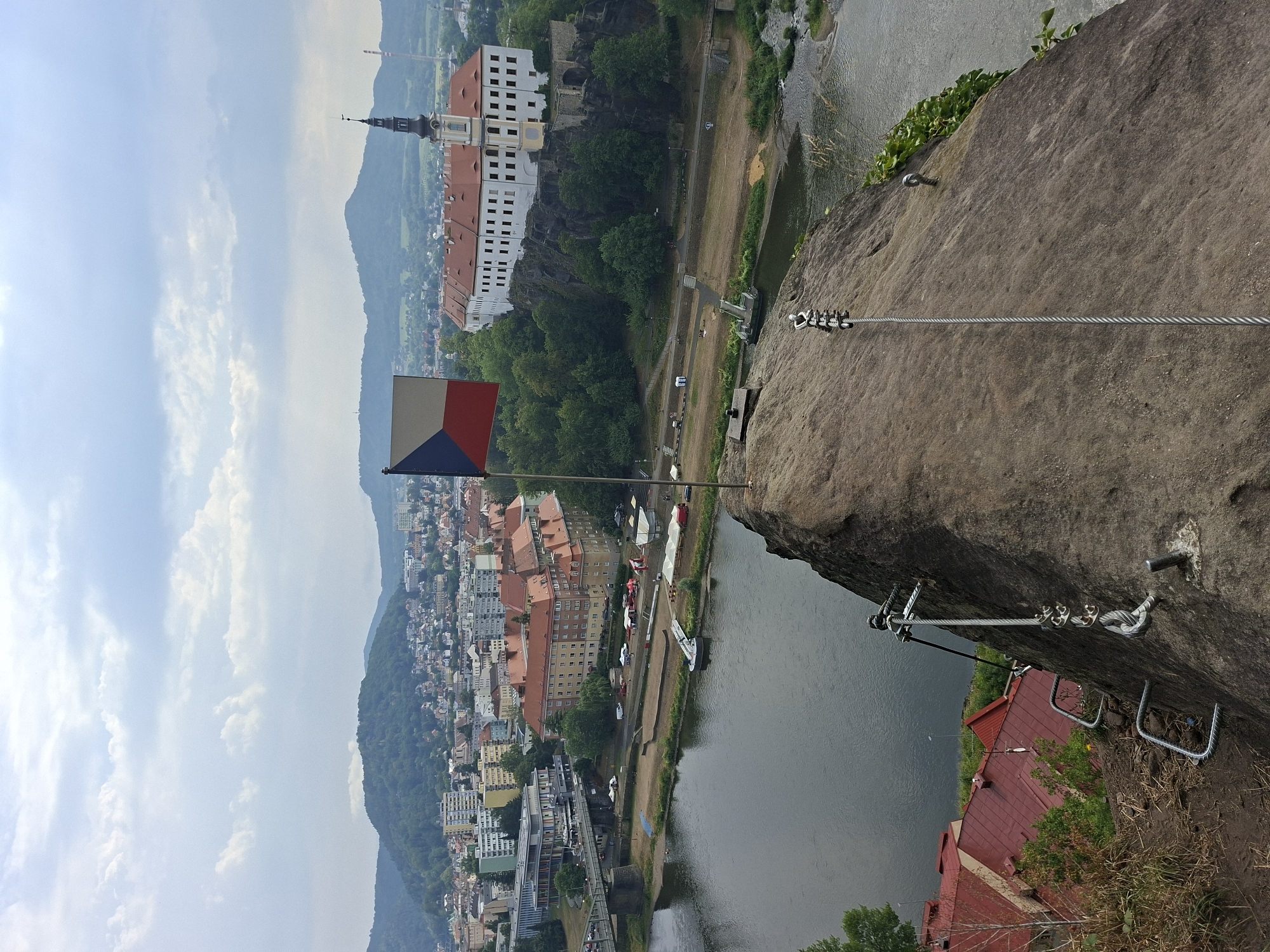 An eye-level, high-angle outdoor shot captures a view of Děčín, Czechia, from a rocky outcrop. In the foreground, a Czech flag flies from a flagpole on the rock. Metal rungs and a cable are visible on the rock face, suggesting a climbing or via ferrata route.
Below, the Elbe River flows, with a sandy bank and several tents set up on it. A small boat or dock is visible in the river. Across the river, a bridge connects to the city, which features a mix of modern and older buildings, including a prominent white building with a red roof and a tower on a rocky hill. Distant green hills and mountains form the backdrop under a cloudy sky. The scene appears to be a scenic overlook or a point of interest for tourists.