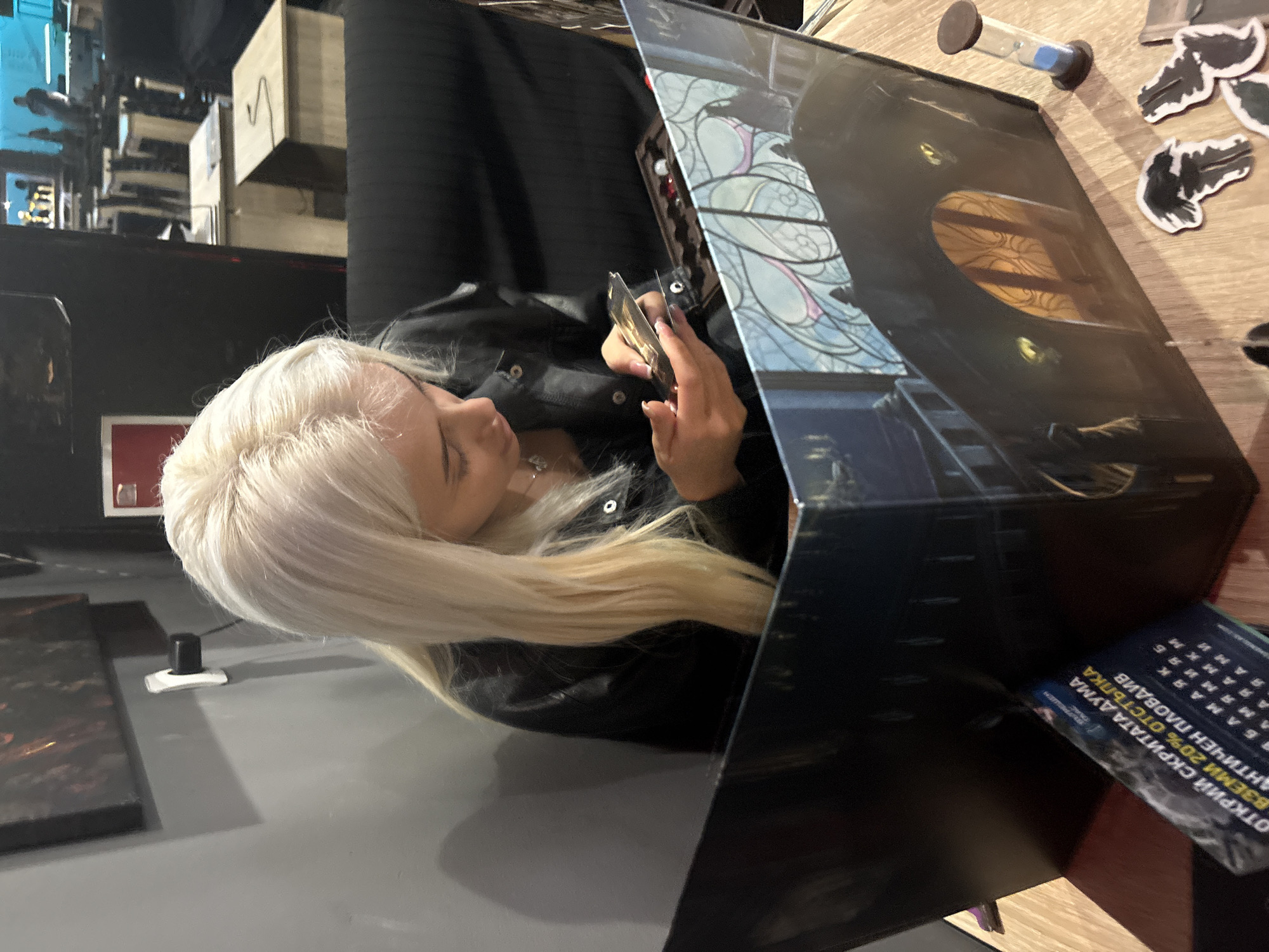 A blonde woman is seated at a table in a room, examining a playing card. A game master's screen stands in front of her. An hourglass and several small black and white tokens are on the table near the top right corner. The location appears to be indoors, likely in Plovdiv, Bulgaria. A poster partially obscured by the game master's screen contains Cyrillic text, a linguistic element indicative of the region.