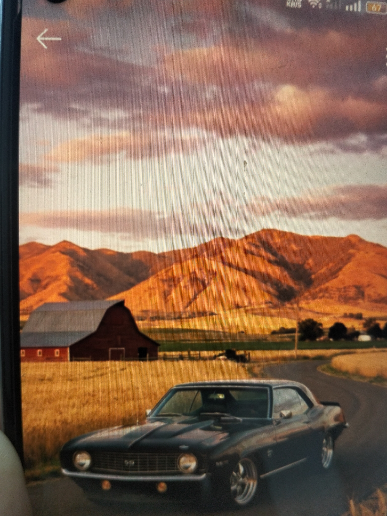 The image depicts a classic black Chevrolet Camaro SS parked on a rural road. The car is facing the viewer, angled slightly to the right. In the background, a red barn with a gray roof is situated in a field of golden wheat. Beyond the field, rolling hills bathed in the warm light of sunset or sunrise dominate the landscape, with a dramatic sky filled with pink and orange clouds. There are no people visible in the image. The scene appears to be set in a countryside environment. The location in the city of Payagaboh, Indonesia is not depicted in this image, as the scene suggests a rural, possibly American, landscape.