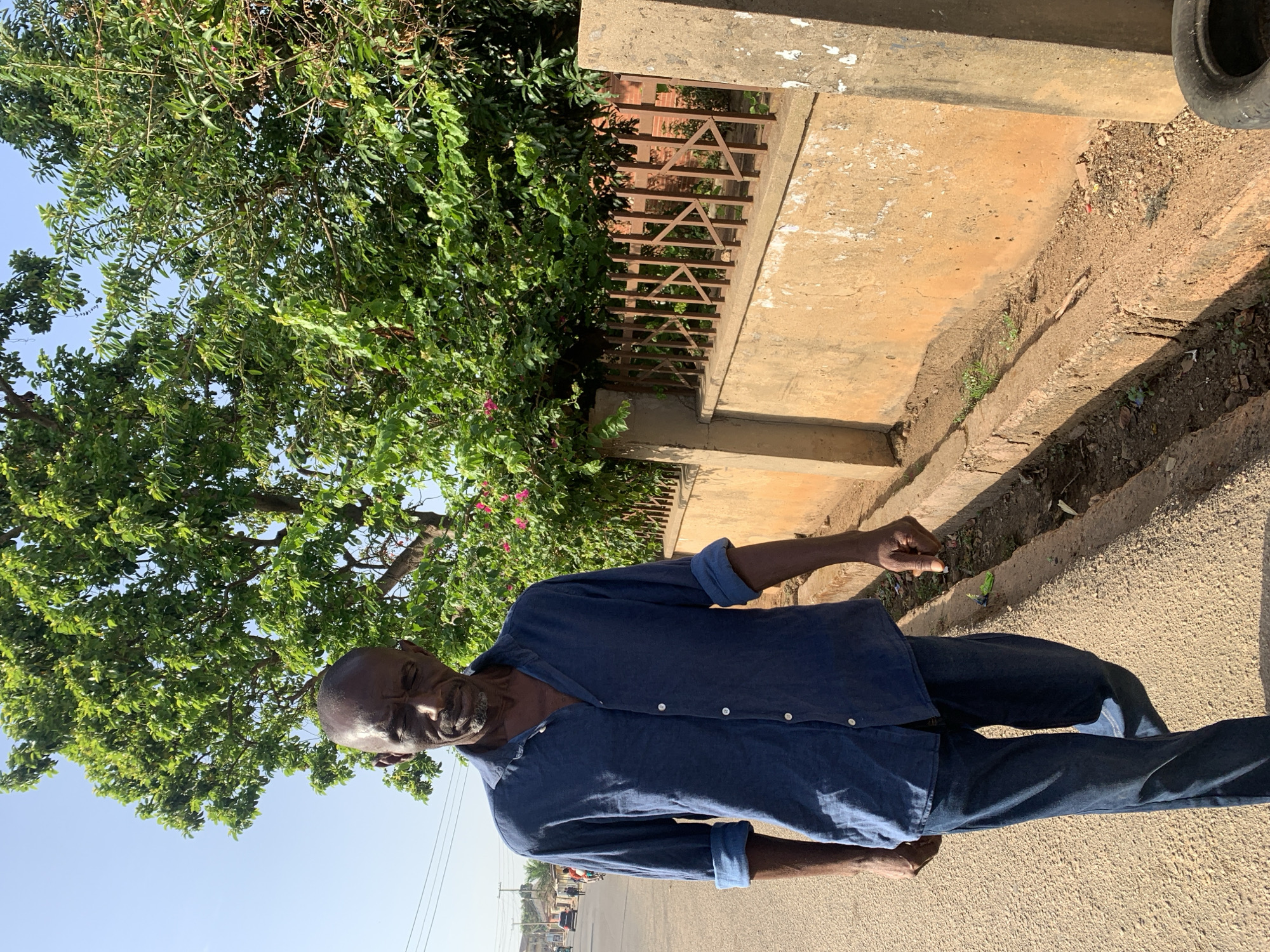 The image captures a man standing on a street in Jos, Nigeria. He is wearing a blue denim shirt and jeans. Behind him is a building with a fence, and a tree with green foliage. In the background, a street view with distant buildings and power lines is visible. The weather appears sunny.
