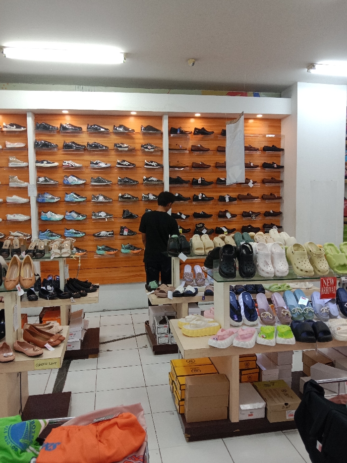 This image depicts the interior of a shoe store in Langenharjo, Indonesia. A prominent wooden-paneled wall serves as the primary display, fitted with numerous clear acrylic shelves exhibiting a wide assortment of men's and women's footwear, including athletic shoes, casual shoes, and formal options, meticulously arranged in rows. A single male individual, seen from behind, wearing a black t-shirt and dark trousers, stands centered before this wall, appearing to inspect or organize the displayed shoes. In the foreground and midground, multiple light-colored wooden display tables showcase diverse women's footwear, such as ballet flats, pumps, sandals, slides, and platform clogs; some displays include "NEW ARRIVAL" signage. Cardboard shoe boxes are stored beneath these tables. The store features a white tiled floor and is illuminated by overhead fluorescent lighting. In the bottom-left corner, a partial view of a container reveals folded textile items, specifically green and orange garments.