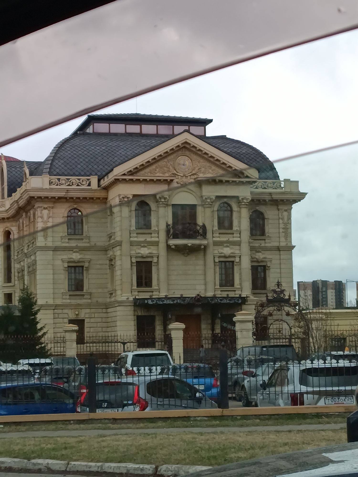 The image shows a three-story building in Krasnoyarsk, Russia. The building has a light-colored facade with decorative architectural elements and a dark domed roof. A balcony is present on the second floor. A wrought iron fence surrounds the property, obscuring a view of several parked cars. In the background, a modern high-rise building is visible. The photograph appears to have been taken from inside a vehicle, through a partially obscured window.