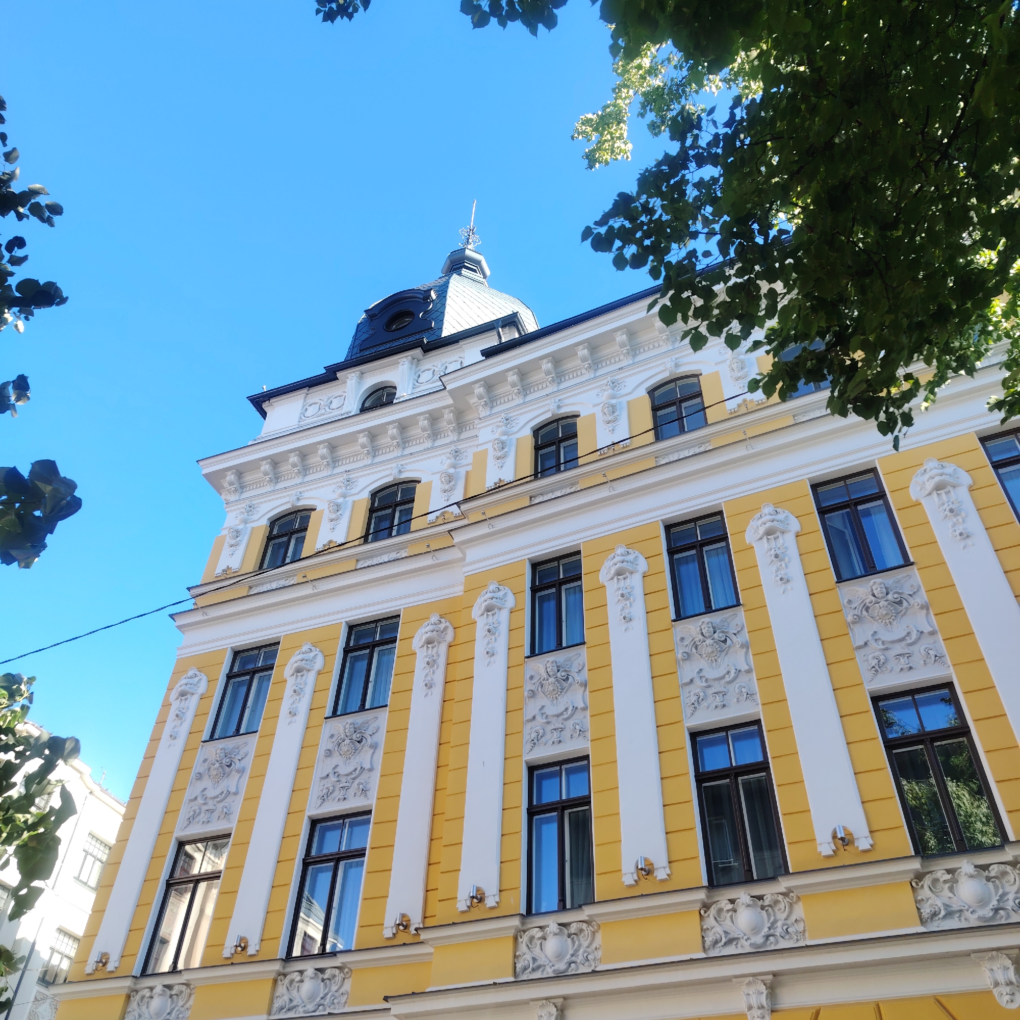 The image depicts a low-angle view of a building facade in Riga, Latvia. The building features a yellow exterior with white decorative elements, including ornate columns and sculptures flanking the windows. The windows are dark with brown frames. A black dome-shaped roof tops the building, with a spire extending upwards. The sky is a clear, bright blue. Tree foliage is visible in the corners of the frame, partly obscuring the building and sky.