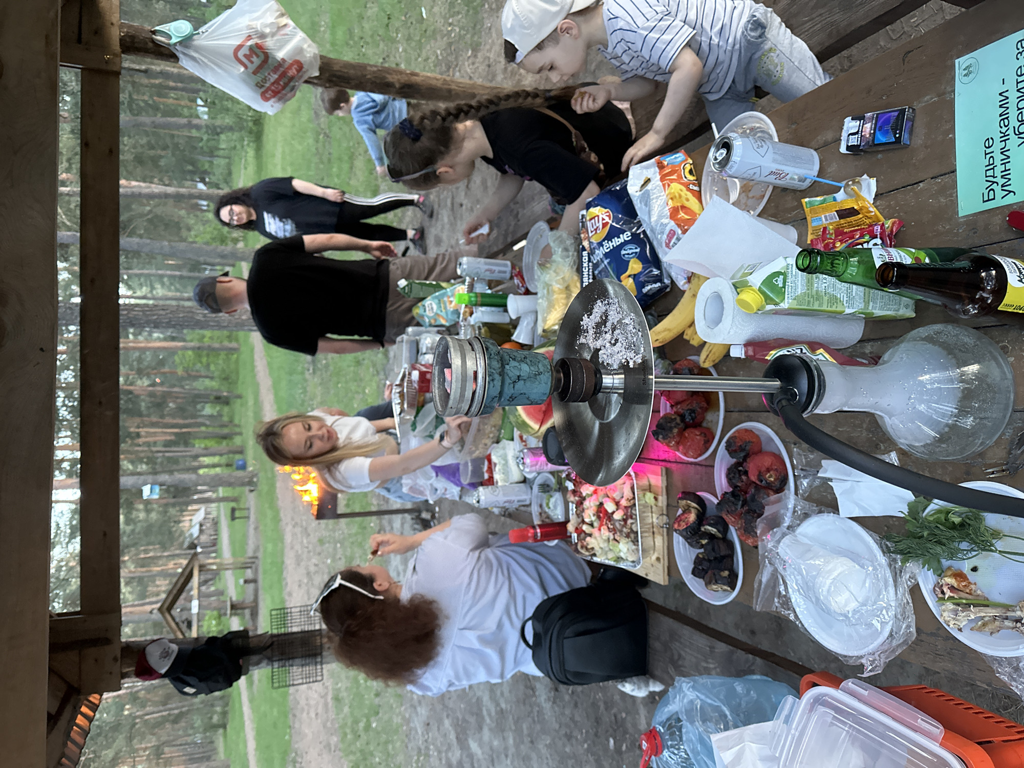 A group of people are gathered around a wooden picnic table in a forested outdoor setting. A hookah is placed prominently in the center of the table. Food items like watermelon, tomatoes, salads, and roasted eggplant are scattered across the table, along with snack foods like chips and bananas. Beer bottles and juice cartons are also visible, along with paper plates and empty plastic containers. Some of the people are interacting with the food and hookah, while others appear to be standing or walking nearby. A bonfire is burning in the background. A plastic bag hangs from a tree. The area appears to be a wooded recreational area. A sign written in Russian is partially visible, but difficult to read. The location and time of year cannot be determined.