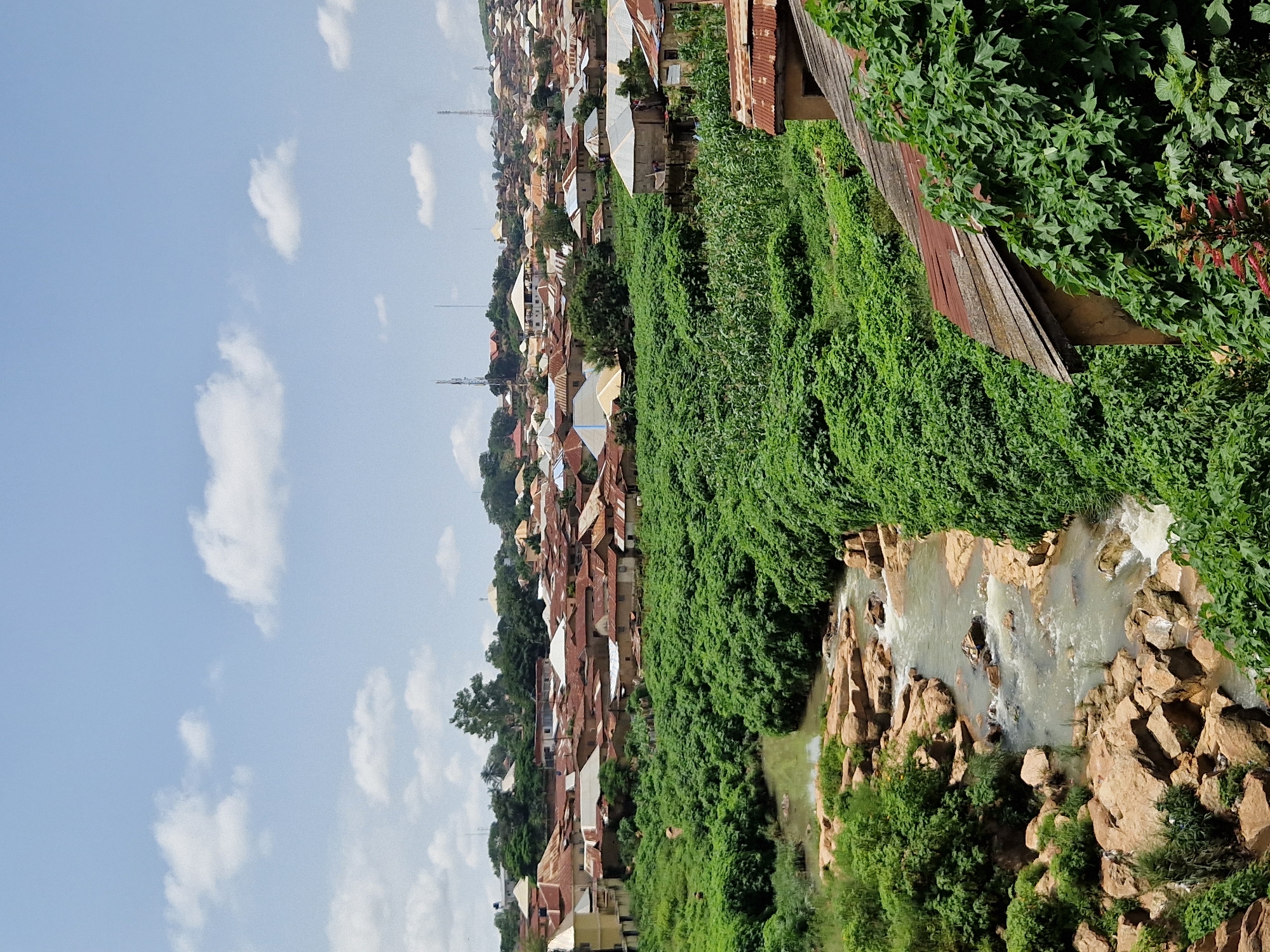 The image captures a wide-angle view of a residential area in Jos, Nigeria, characterized by a densely packed collection of buildings with predominantly red, corrugated iron roofs. The settlement is situated on a series of undulating hills, with a natural landscape of rocky terrain and lush green vegetation that includes a small river flowing through the foreground. The river has a shallow, rocky bed, with visible water flowing over stones and creating small cascades.
The residential structures are varied in size and architectural style, with some appearing older and more weathered than others. Many buildings are two-story, constructed with cement or brick and topped with the characteristic metal roofs. In the midground and background, the houses blend into the hillside, with more distant structures becoming less distinct. Tall telecommunication towers are visible on the skyline above the town, indicating the presence of modern infrastructure.
The foreground is dominated by dense green foliage that grows prolifically along the banks of the river and up the slopes. Some areas show evidence of agriculture, with rows of corn plants visible amidst the other vegetation. A few trees are interspersed within the urban landscape, adding to the greenery. The sky above is a clear blue with scattered white cumulus clouds. No people are clearly visible in the image, and there are no overt signs of activity or interaction. The overall impression is one of a vibrant, living community integrated with its natural surroundings.