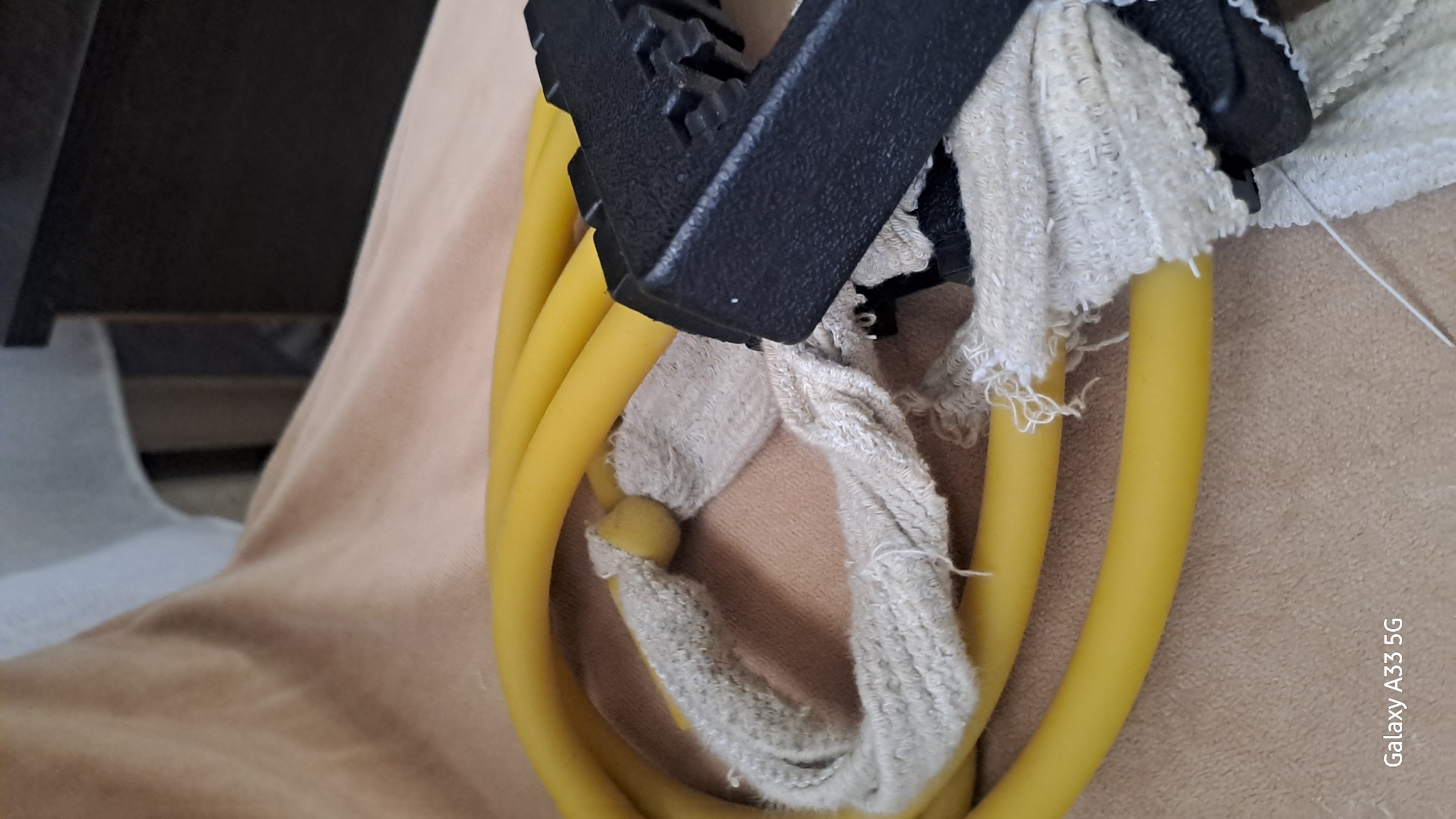 A close-up, upward angled shot shows a yellow resistance band. The band is looped and attached to a black plastic handle. The point where the band attaches to the handle is wrapped with a frayed white cloth. The background consists of a textured, tan surface, possibly a blanket or upholstery, and a blurred dark wooden furniture piece in the upper right corner. A white fabric sheet is visible in the upper left corner. The bottom left corner of the image shows the text "Galaxy A33 5G". The scene appears to be indoors. The location of Ognevka, Kazakhstan, is not discernible from the image.