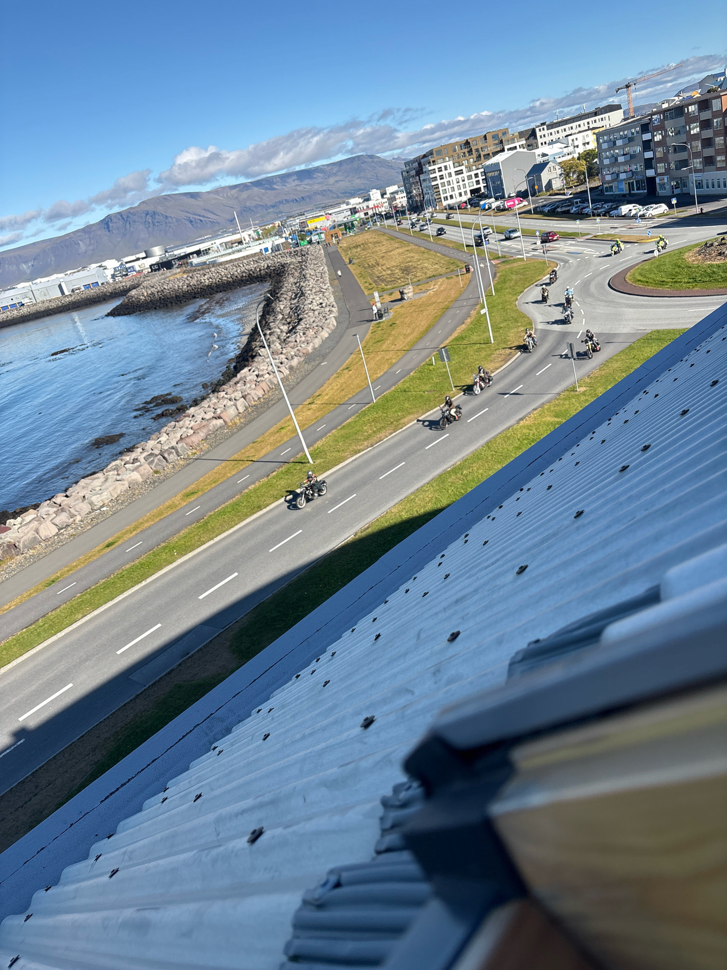 The image shows a wide, scenic view of a coastline in Reykjavik, Iceland. A paved road runs alongside a body of water, with a stone embankment separating the road from the sea. Several motorcyclists are riding on the road, some in a group and others individually. In the background, there are buildings, mountains, and a crane, suggesting an urban or semi-urban environment. The sky is clear and blue with some scattered clouds. The foreground of the image is dominated by a ribbed metal roof, angled diagonally and filling the lower portion of the frame. The perspective suggests the photo was taken from a high vantage point, looking down and across the landscape.