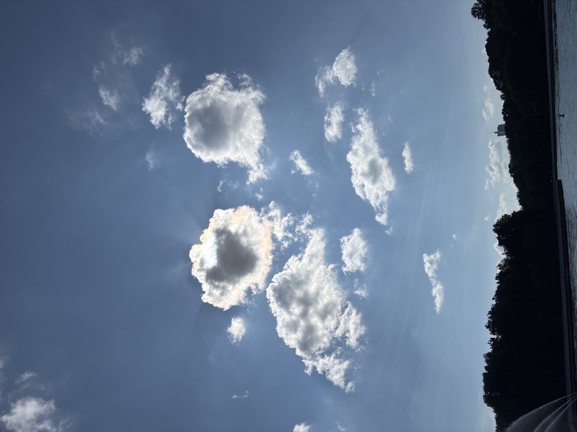 The image captures a bright, sunny day over a body of water, likely a reservoir or river, in Minsk, Belarus. The sky is a vivid blue, dotted with fluffy white clouds. One prominent cloud formation in the upper center of the frame is partially illuminated by the sun, creating a prismatic effect with iridescent colors, indicating it might be a nacreous cloud. In the foreground, the dark silhouette of a dense line of trees forms a border along the water's edge, suggesting a park or natural area. A faint structure, possibly a building or tower, is visible on the horizon, peeking above the treeline. A fountain sprays water on the left side of the image, adding a dynamic element to the scene. The water itself appears calm and reflects the sky and trees. The overall atmosphere is serene and picturesque.