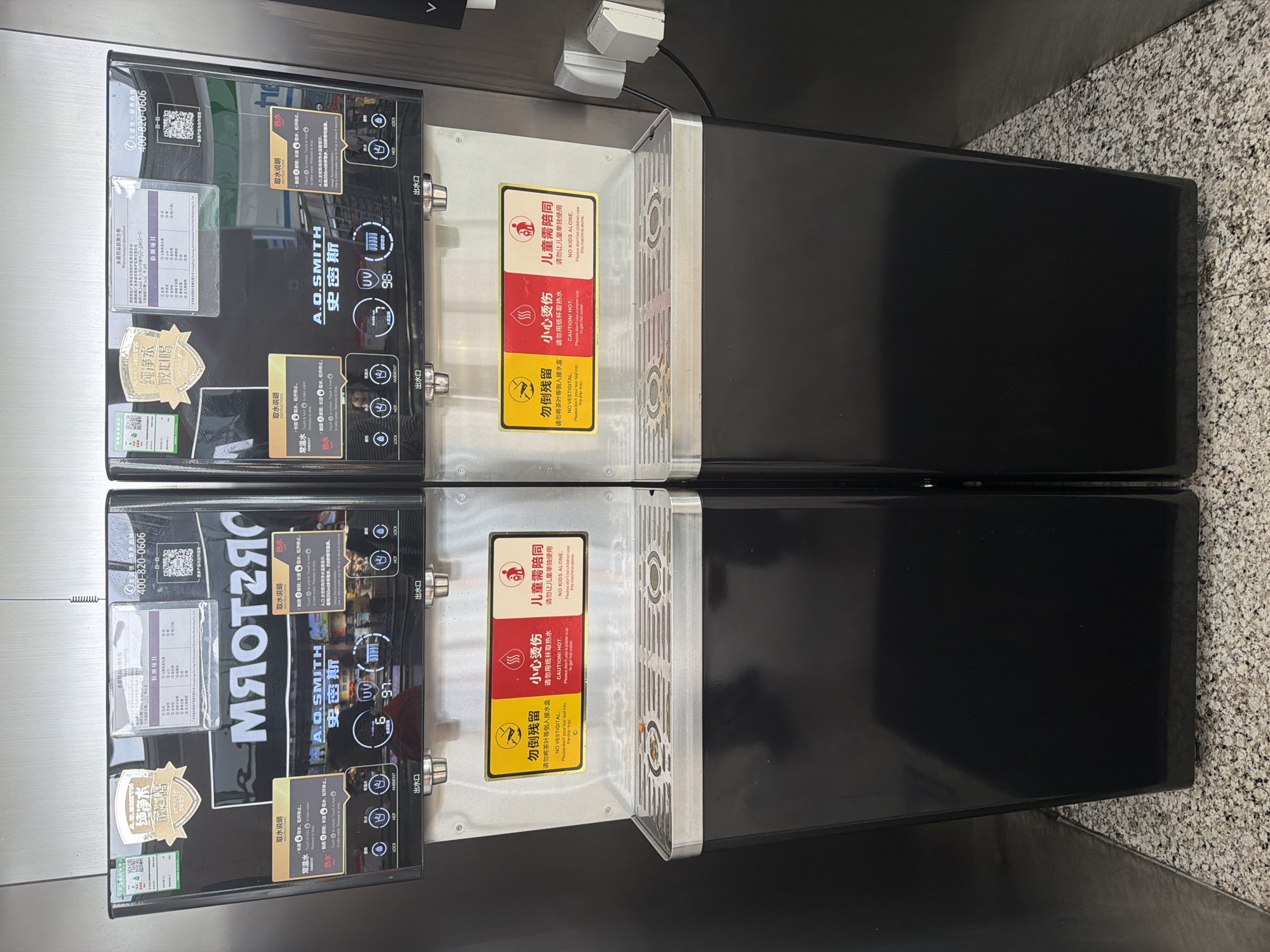 Two identical A.O. Smith water dispensers are displayed side-by-side in what appears to be an indoor commercial or public setting. The dispensers have black front panels with digital displays showing "UV" and numbers like "98." and "97.", likely indicating water quality or temperature. Each unit features two spouts, one for ambient water and one for hot water, along with buttons for "LOCK," "HOT," and "AMBIENT," and an "AMOUNT" indicator. Below the spouts, each dispenser has a stainless steel drip tray with a grated surface.
Prominently displayed on each dispenser are several labels and warnings. Instructions for dispensing water ("取水说明") are provided for both ambient and hot water, indicating users should press and hold to dispense and release to stop, with an additional "unlock" step for hot water. A "warm reminder" suggests using a 350ml cup for hot water, which fills in approximately 8 seconds.
Three main warning signs, in both Chinese and English, are visible on the drip tray area: a red sign cautioning "CAUTION! HOT." and advising against using paper cups for hot water; a yellow sign stating "NO VESTIGITAL." and asking users not to pour tea leaves into the drip tray; and another red sign warning "NO KIDS ALONE." and instructing not to let children use the machine unsupervised.
Other visible text includes the brand "A.O. SMITH" (史密斯), a service hotline "400-820-0606," and a "Water Sample Qualified" (水质样品检测合格) report with inspection items ("检测项目") and the phrase "Pure water, drink with confidence" (纯净水 放心喝). QR codes are also present on the front panels. The background features a dark wall and a light-colored, speckled countertop or floor. No human activity is depicted.