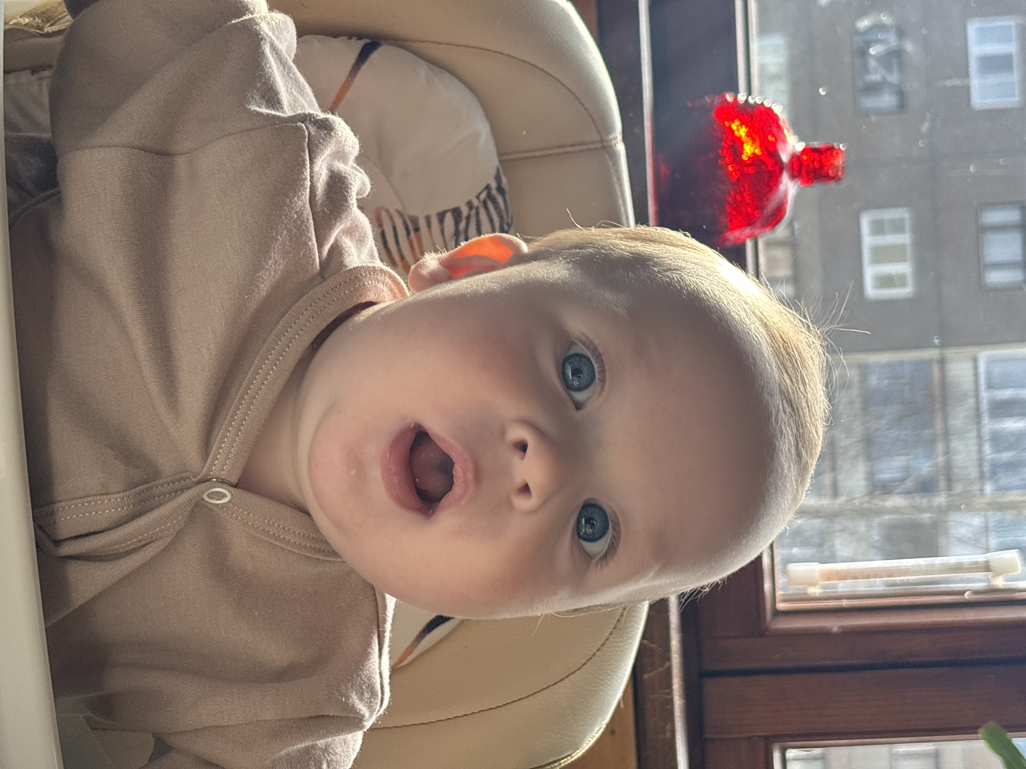 A bright, close-up portrait shows an infant or young toddler with fair skin, light blonde hair, and wide, captivating blue eyes, looking directly at the viewer with an open mouth, as if in surprise or vocalizing. The baby is wearing a plain, long-sleeved light beige or tan onesie with a visible snap closure.
The setting appears to be indoors, with the baby seated in a light-colored high chair, parts of which are visible behind and below the child. To the baby's right, a window with a dark brown wooden frame reveals a blurred urban or residential background, consisting of multi-story buildings with numerous windows. Bright daylight illuminates the scene, suggesting a clear, sunny day. A striking, intensely reflective red object, possibly a glittery or sequined ornament, is visible near the window, catching and scattering sunlight. No visible text is present in the image. The scene conveys a sense of innocent curiosity and engagement from the child. The provided location context is Марковский, Russia.