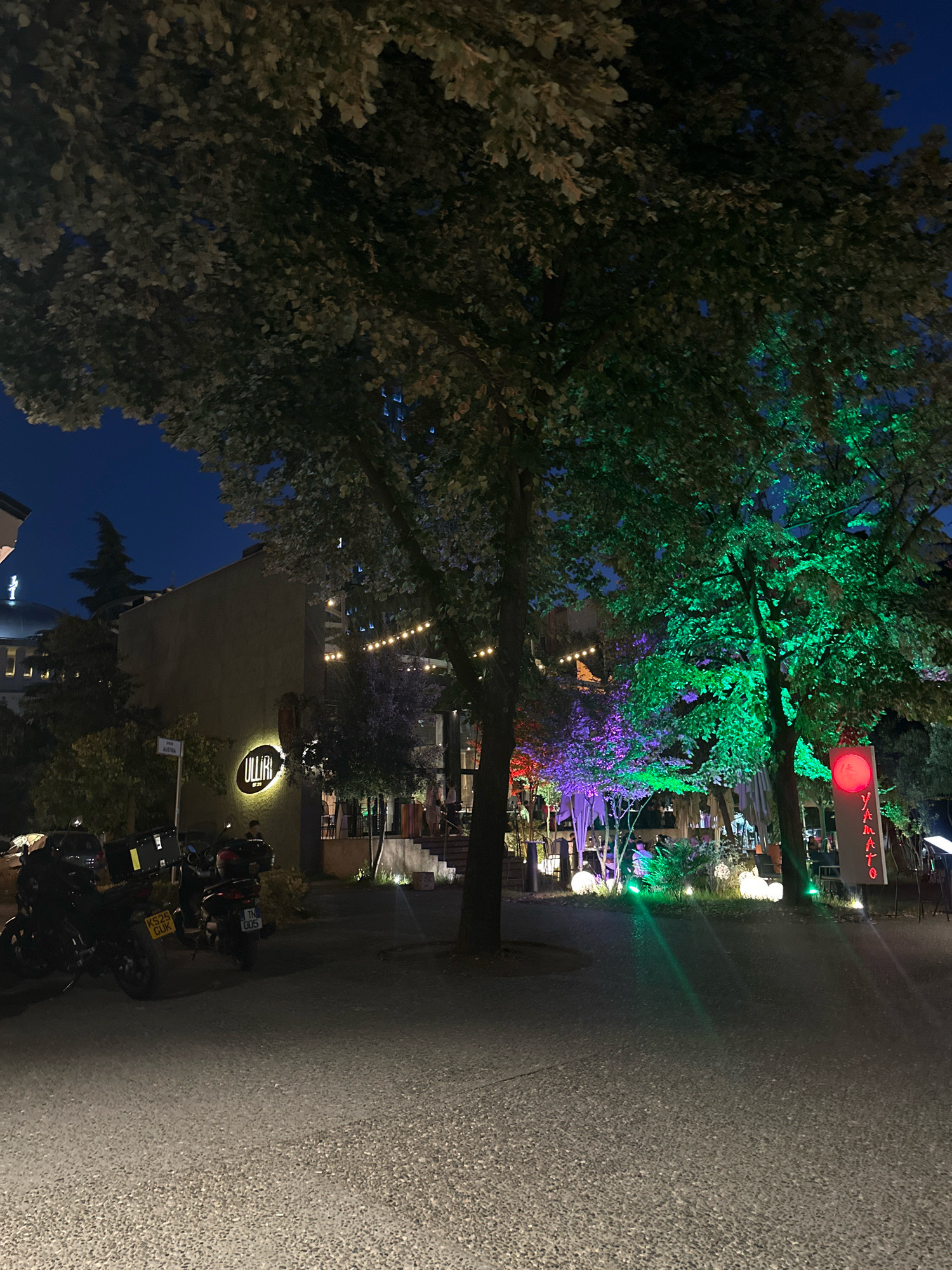 The image depicts an outdoor scene at night in the city of Tirana, Albania. The setting appears to be a street or plaza area adjacent to a building housing a restaurant or bar, indicated by the glowing sign "ULLIRI" and the presence of people seated at tables and illuminated string lights. Several trees are prominent in the foreground and midground, with some of them illuminated by colored lights, specifically green and purple. To the right, a vertical sign with red lighting and the word "YAMATO" is visible, suggesting a Japanese restaurant or establishment. In the left foreground, multiple motorcycles are parked. The ground surface is paved with small stones or pebbles. The sky is dark, indicating nighttime.